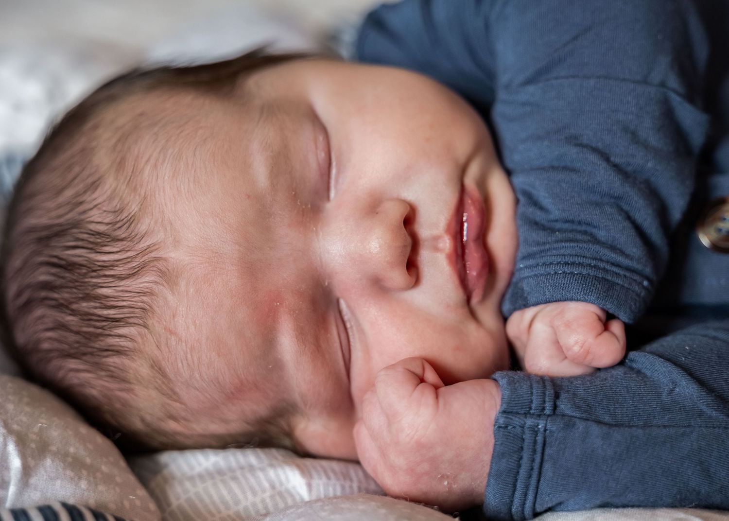 A peaceful newborn baby in a blue outfit sleeps soundly with tiny fist raised near their cheek.