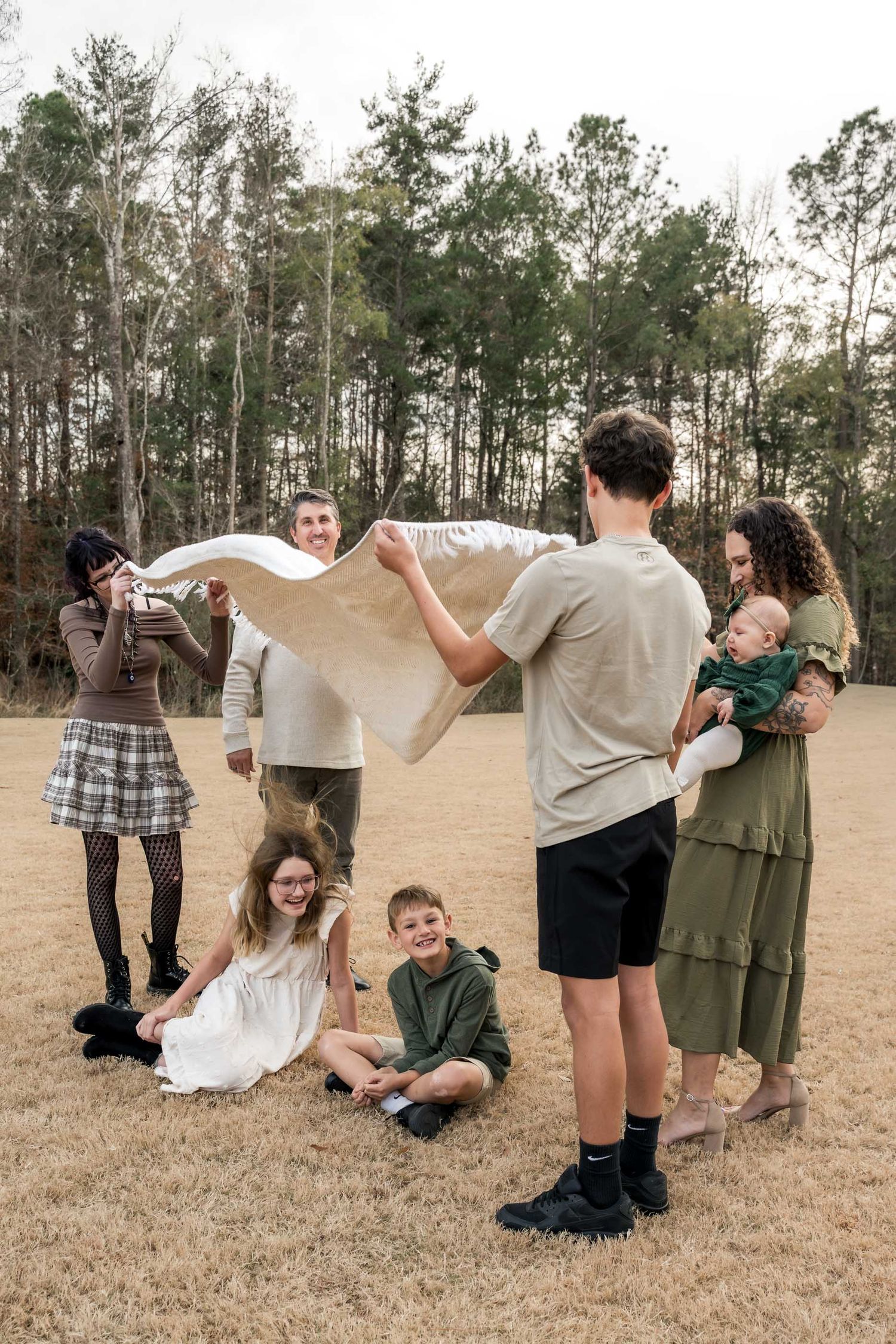 Family gathering outdoors on dry grass with tall pine trees, sharing a blanket while posing for a casual photo session.