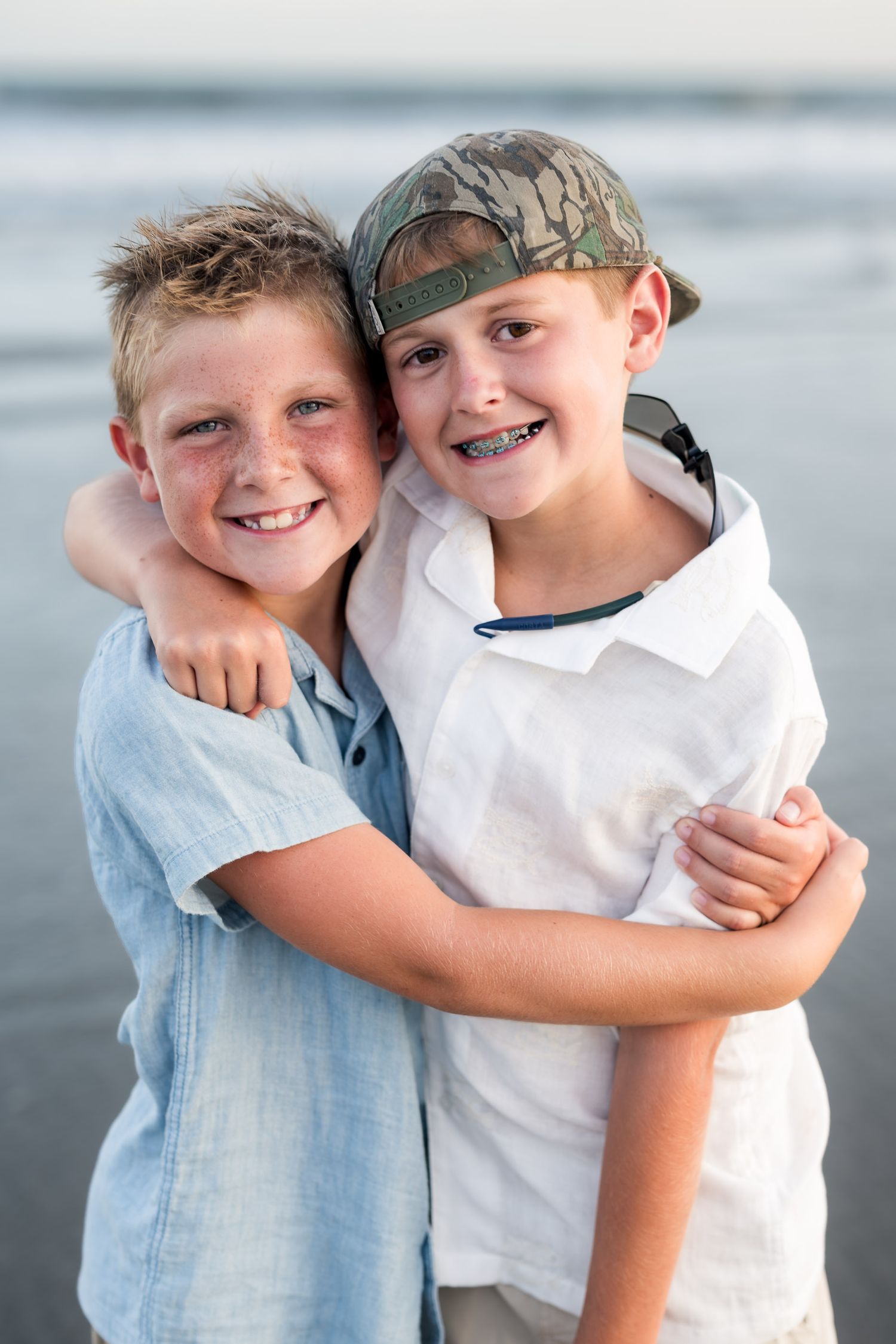Two smiling boys hugging each other on a beach, one wearing a camo cap and braces, the other in a blue shirt.