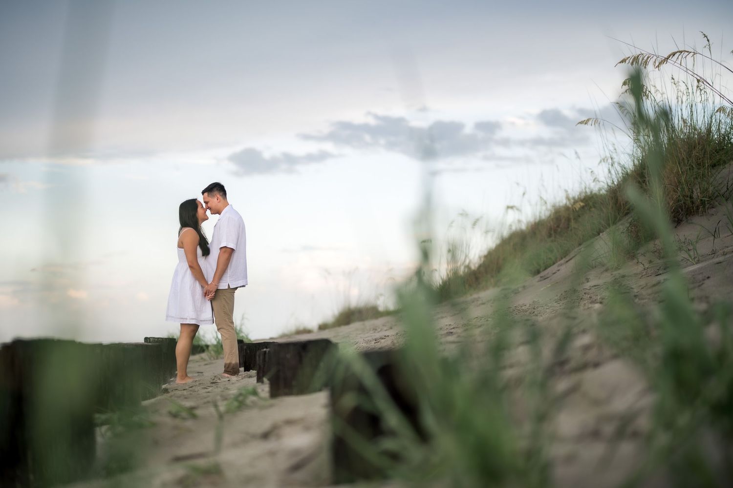 Couple holding hands on sandy beach dunes at sunset, dressed in white, with sea grass and cloudy sky backdrop.