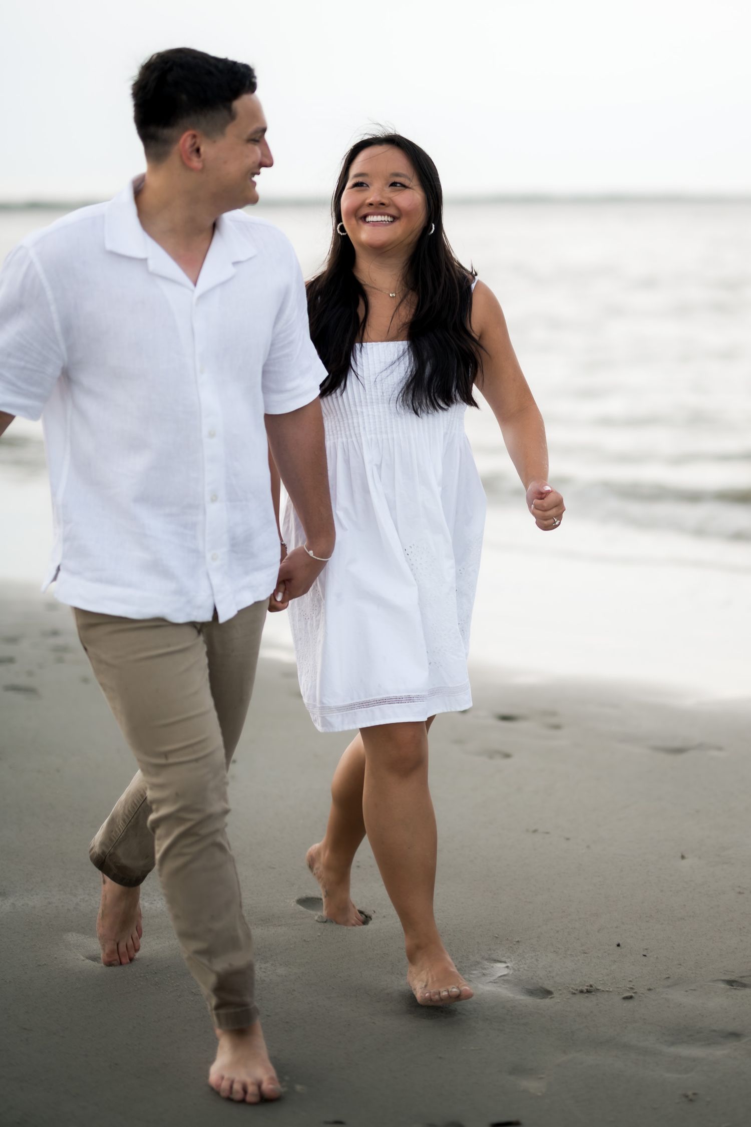 Couple in white outfits walking barefoot on a beach, holding hands and smiling joyfully.
