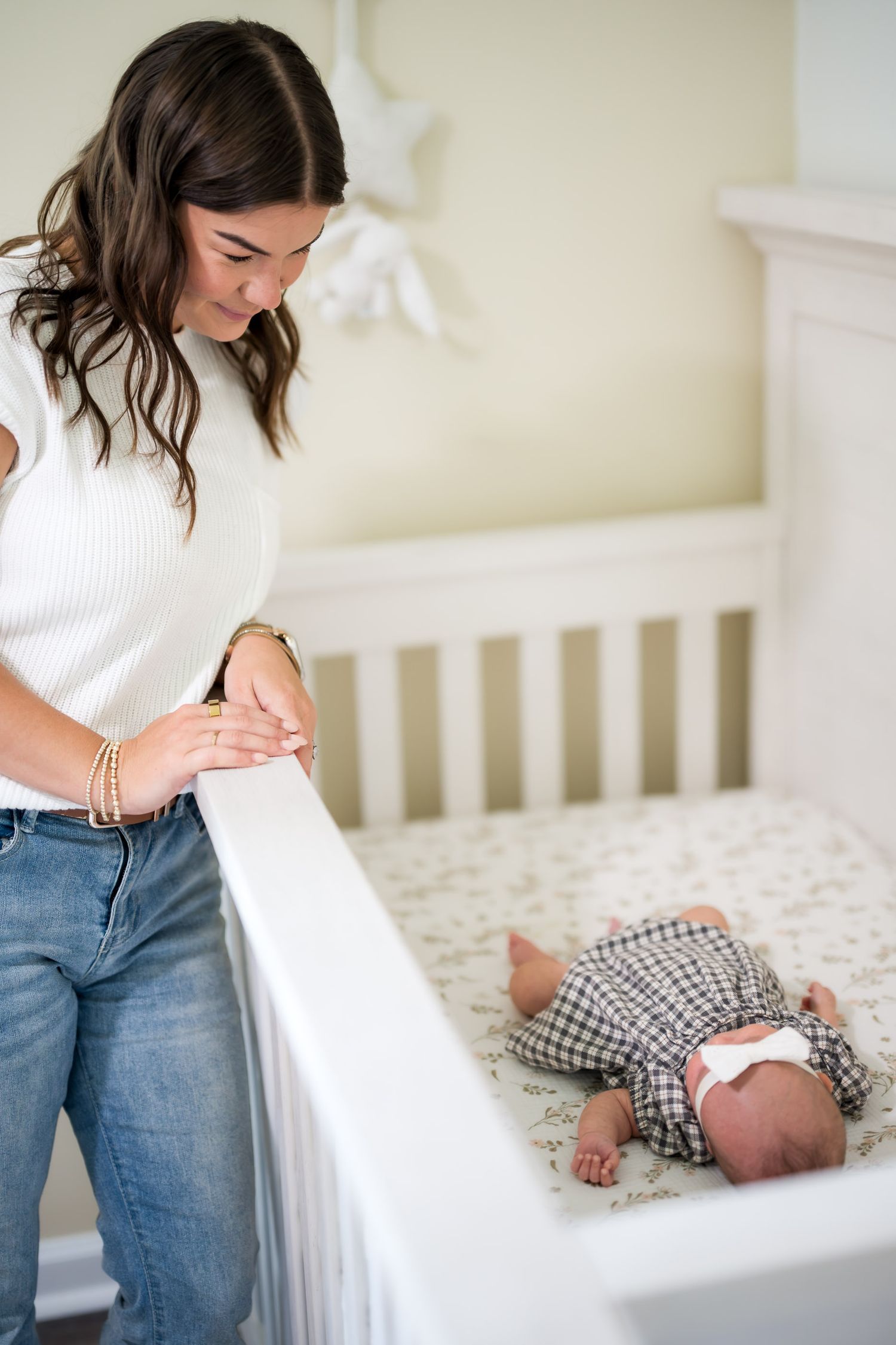 A young mother in a white top and jeans lovingly gazes at her newborn baby lying in a white crib.