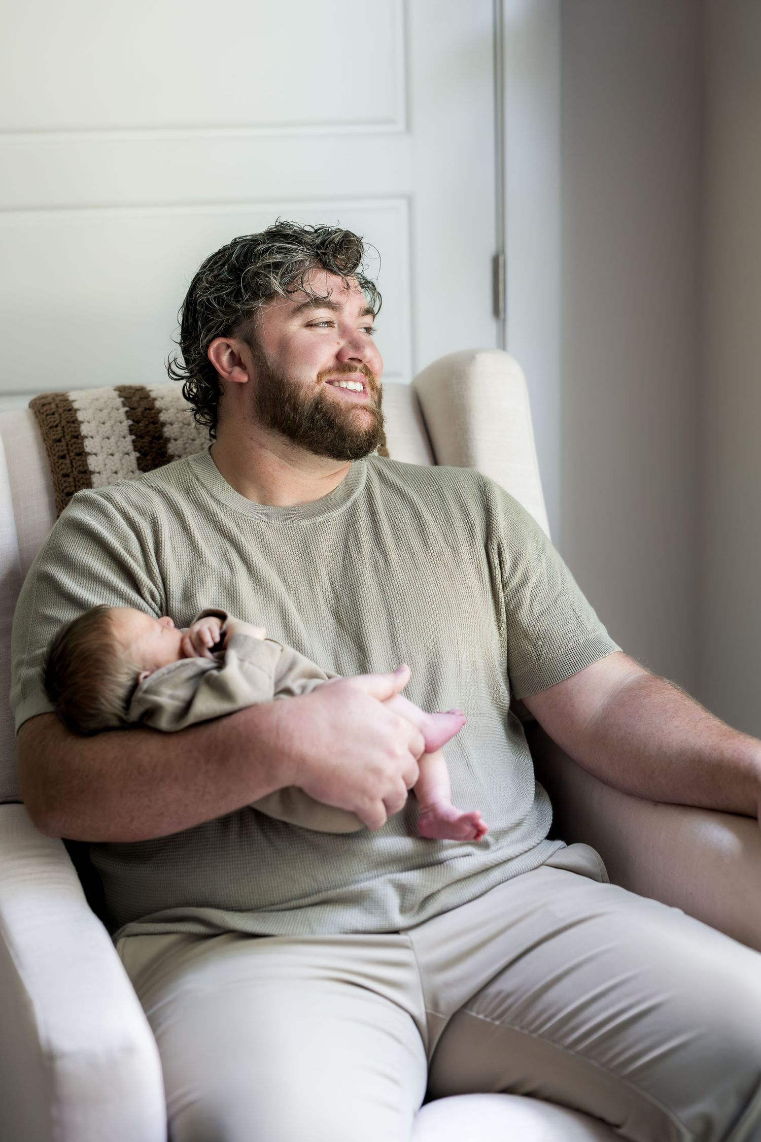Bearded man in olive t-shirt sitting in armchair holding sleeping newborn baby, gazing out window contentedly.