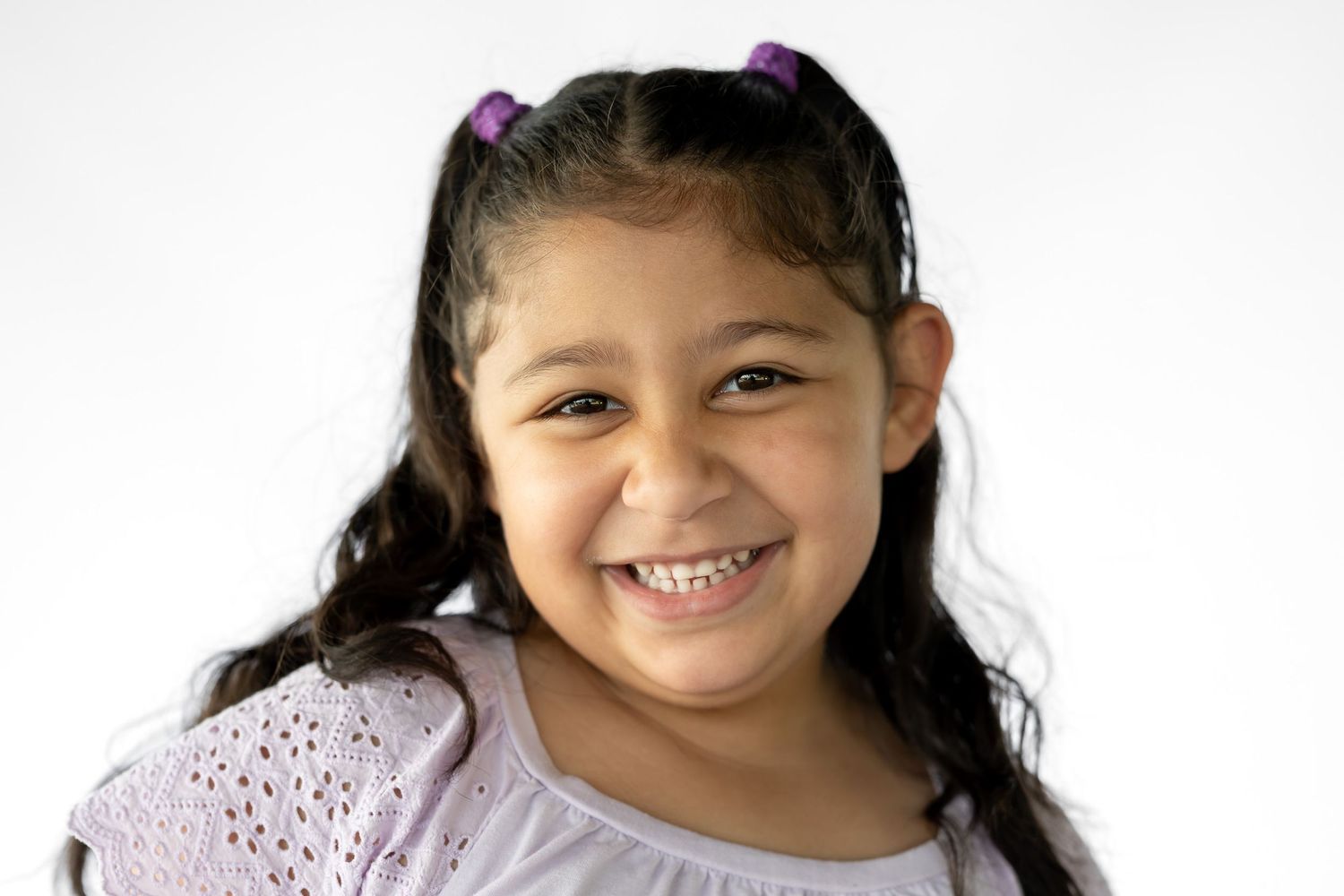 Smiling young girl with dark hair in pigtails wearing a white top against a light background.