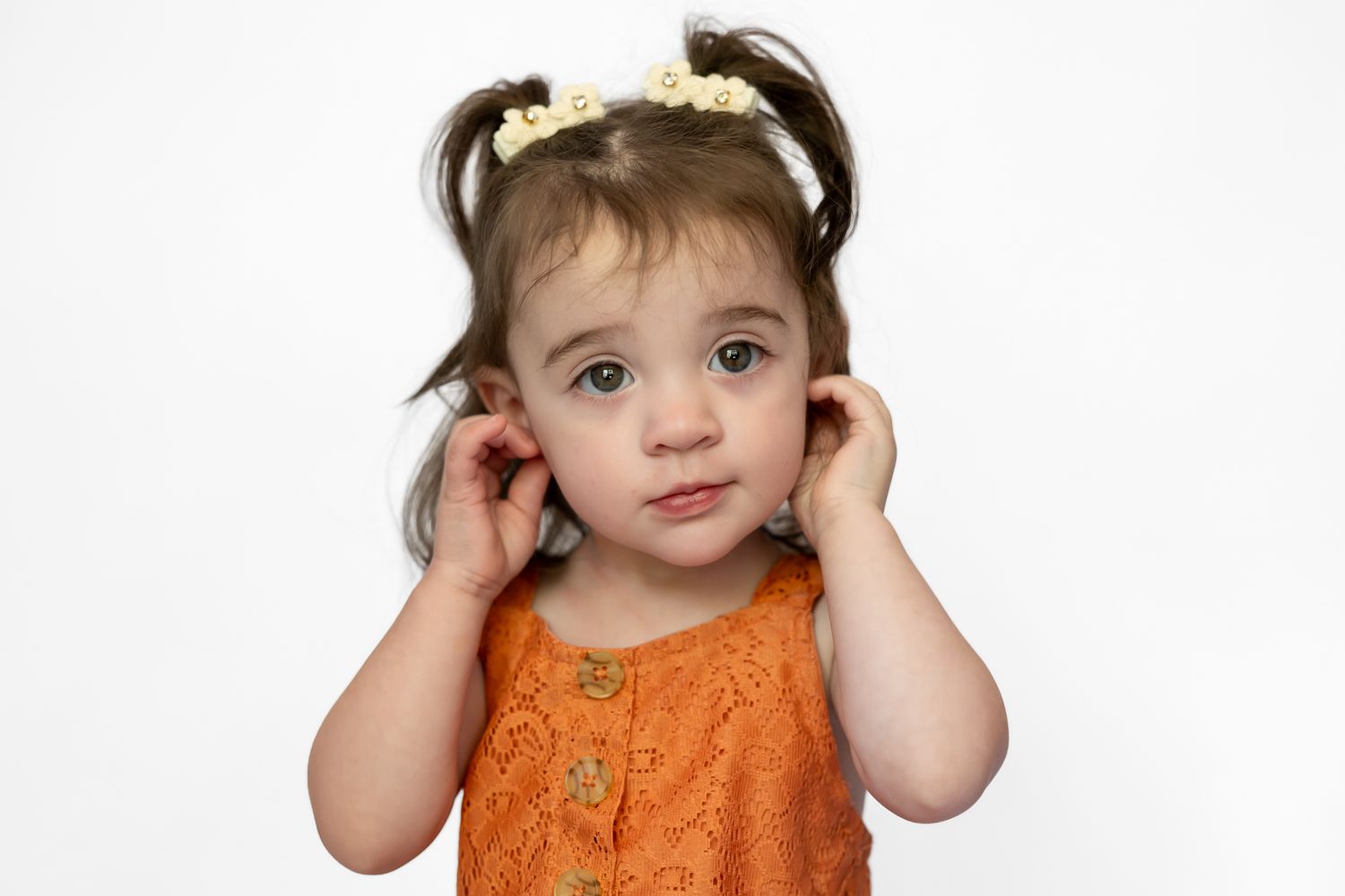 Cute toddler girl in orange lace dress with pigtails and floral headband holding her ears on white background.