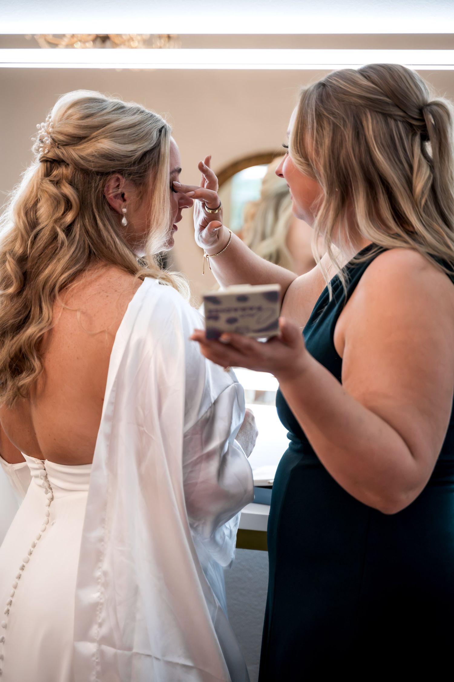 Bridesmaid in green dress applying makeup to blonde bride during wedding day preparation.