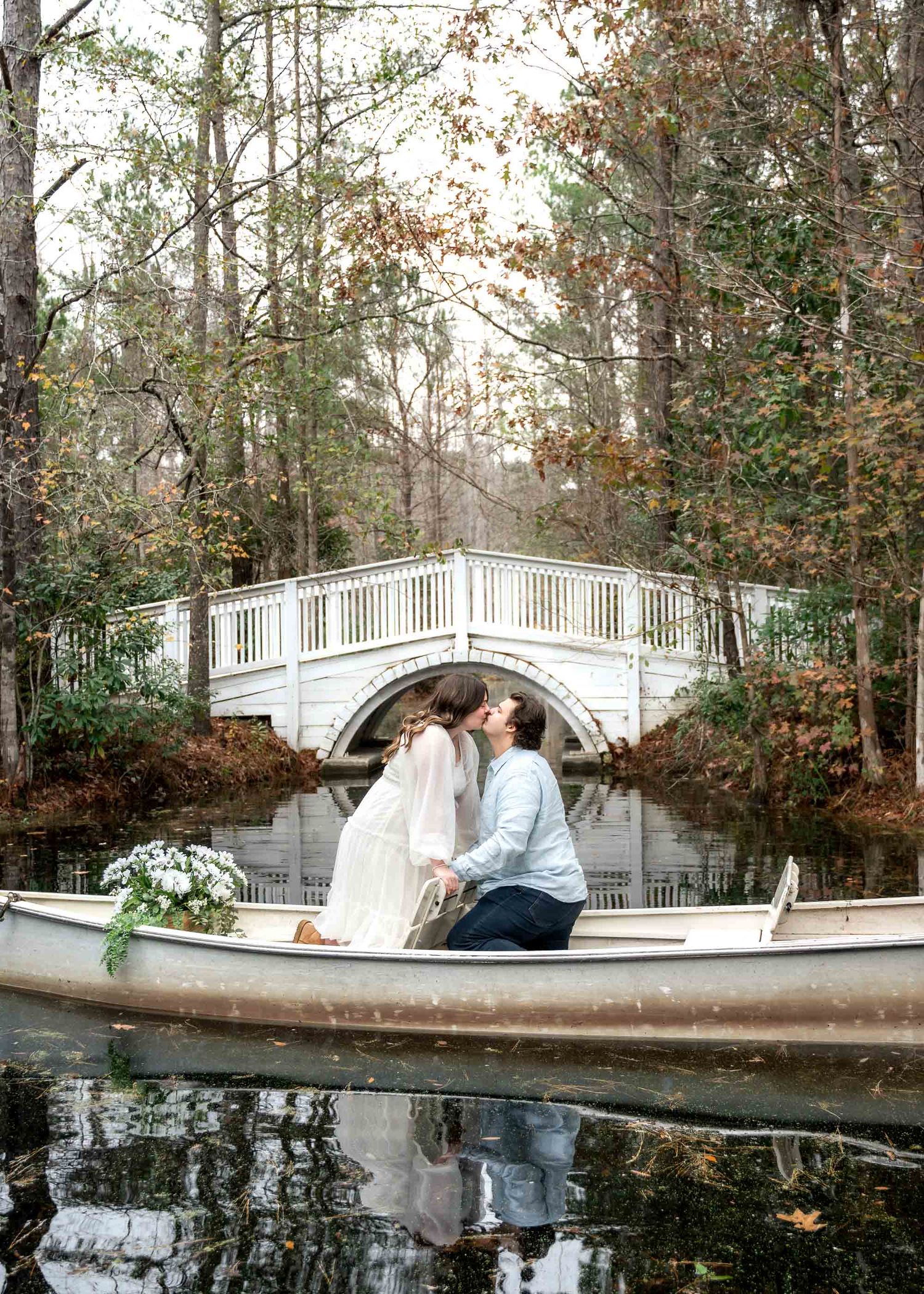 Cypress Gardens. Couple shares romantic kiss in a canoe on a serene pond with a white arched bridge surrounded by autumn trees.