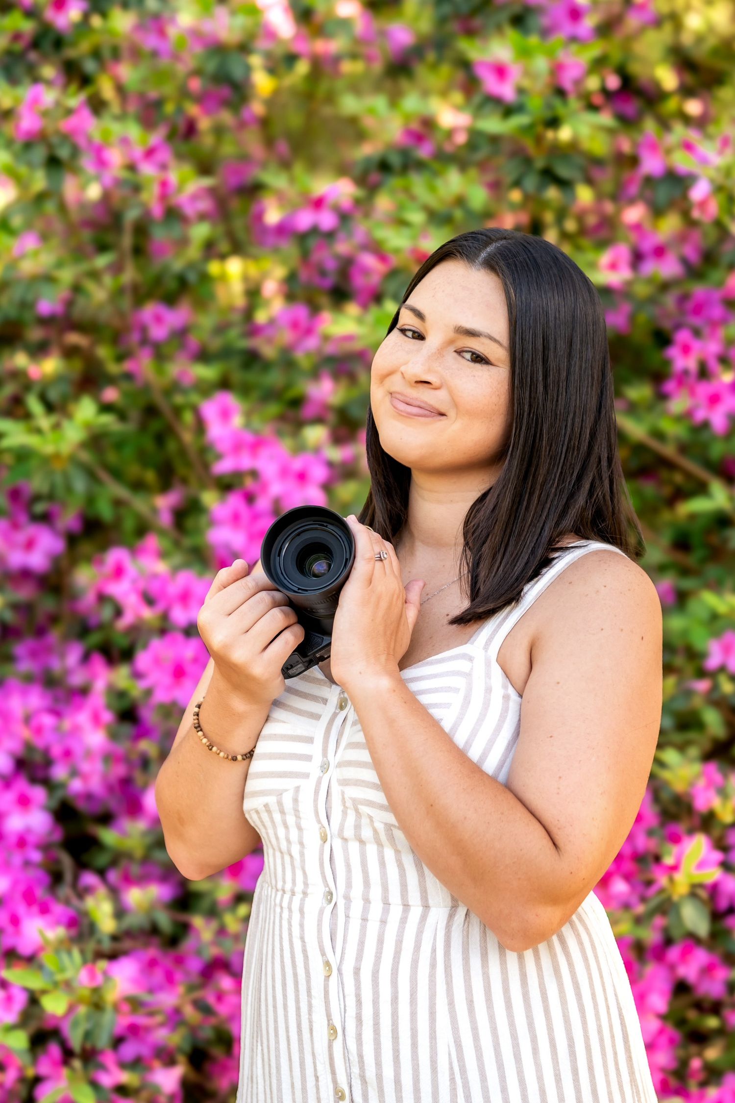 Smiling female photographer holding Nikon camera in front of vibrant pink flowering bushes.