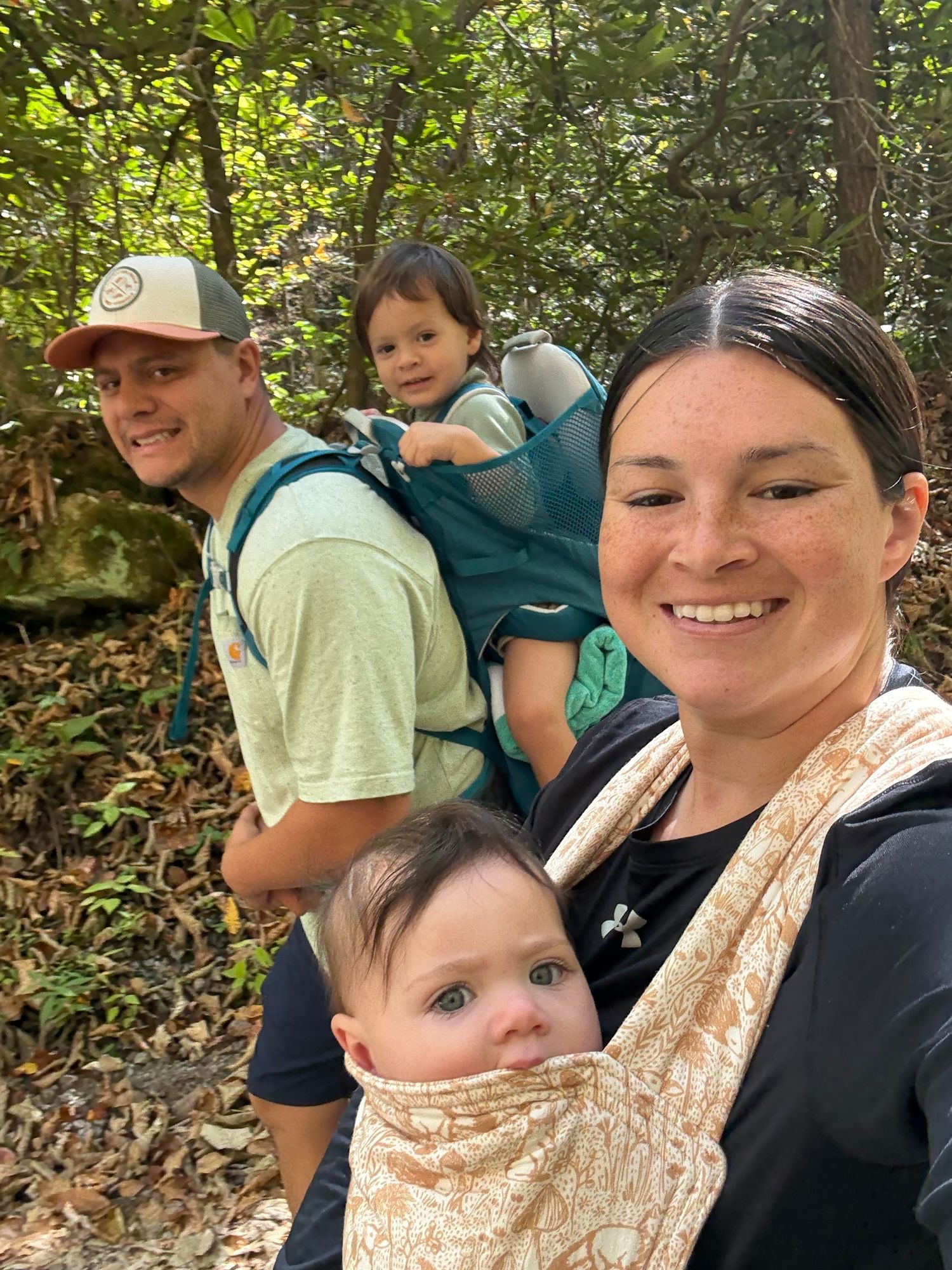 Family of four hiking on wooded trail with children carried in backpack and front baby carrier.