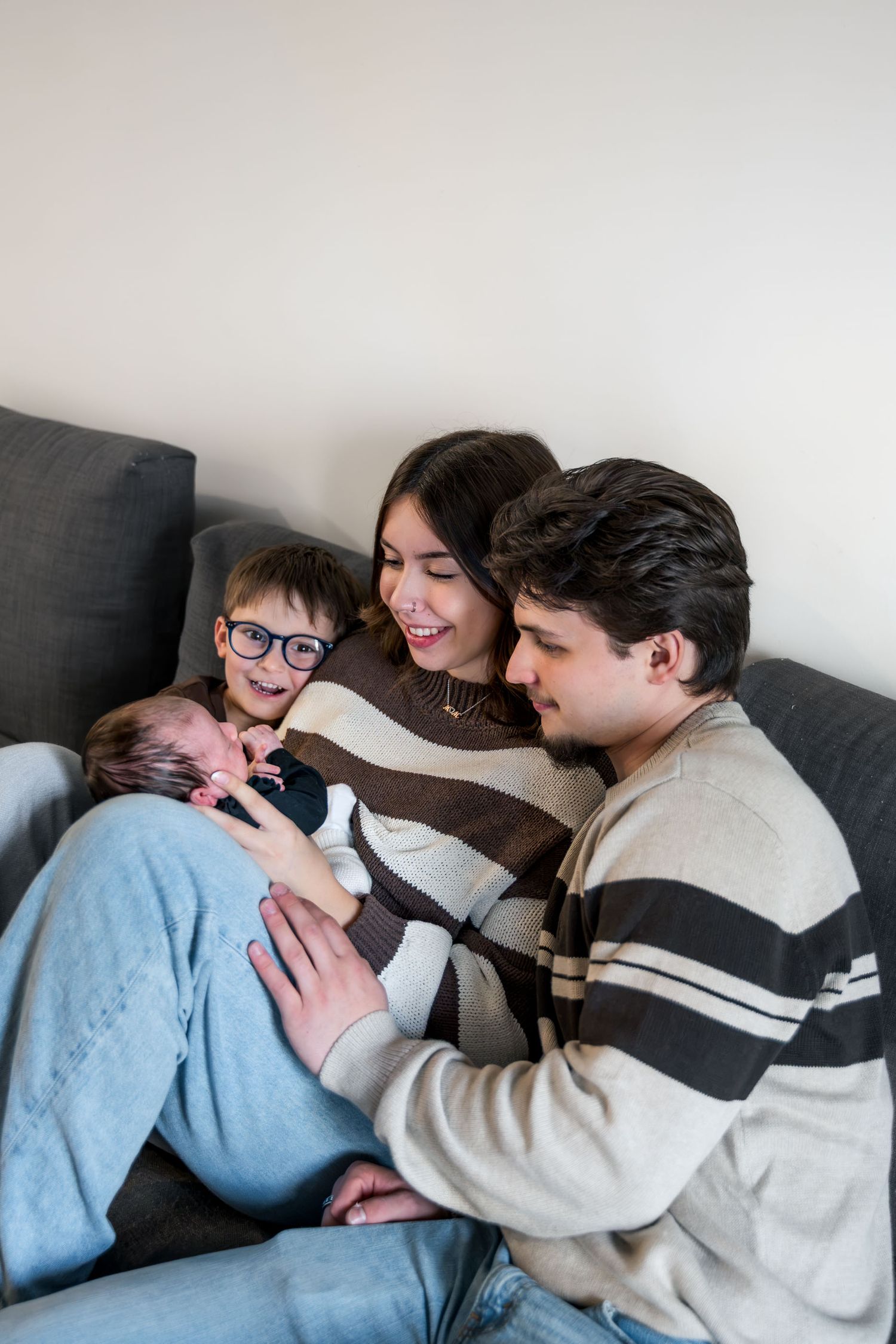 Parents and young boy with glasses smiling at newborn baby on gray couch together.