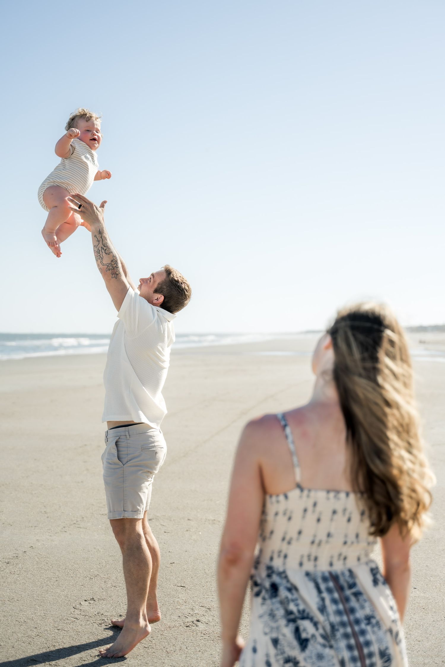 A tattooed father joyfully tosses his laughing baby in the air on a sunny beach while mother watches.