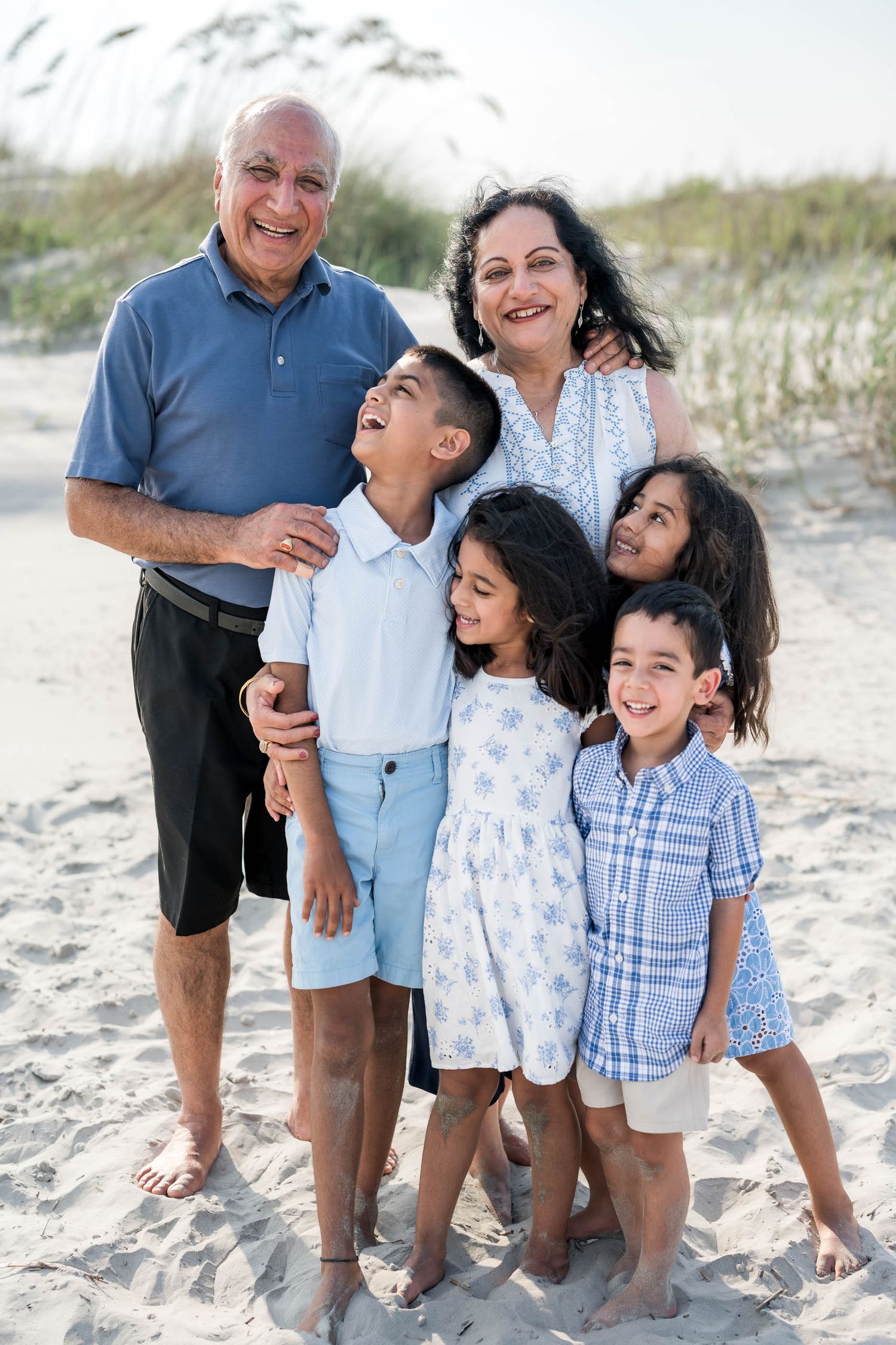 Grandparents smiling with three young grandchildren on a sandy beach, wearing light casual summer outfits.