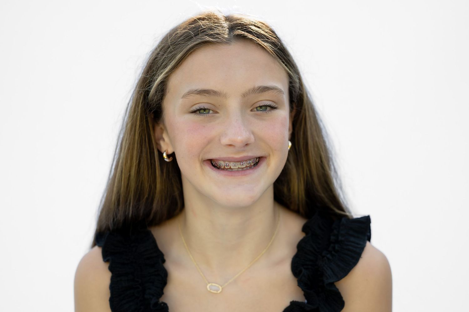 Smiling teenage girl with braces and long brown hair wearing a black top and silver necklace against white background.