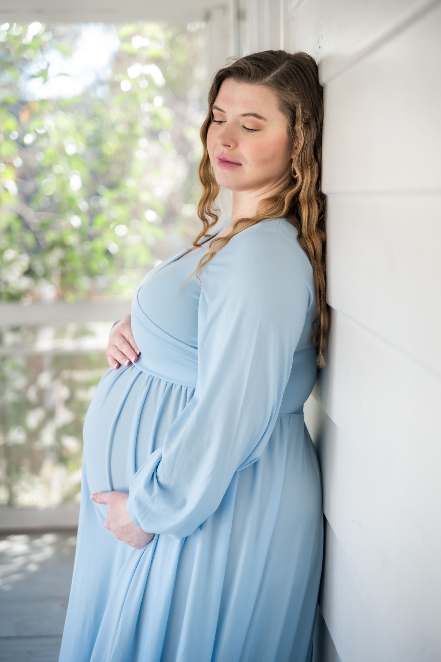 Moncks Corner.Pregnant woman in soft blue maternity gown leans against white wall with eyes closed cradling her baby bump.