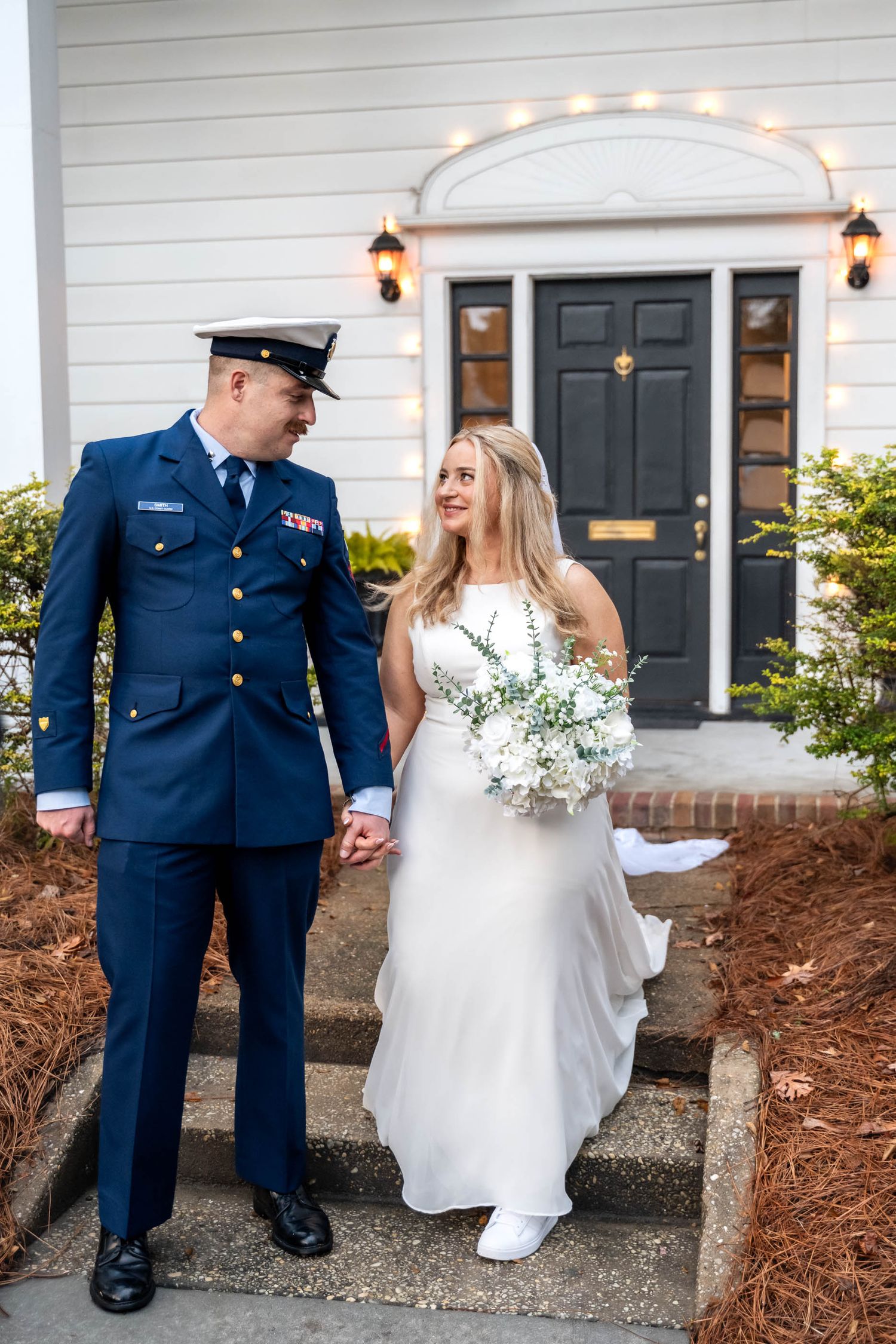 Coast Guard groom in dress uniform and bride holding white bouquet pose on steps outside venue.