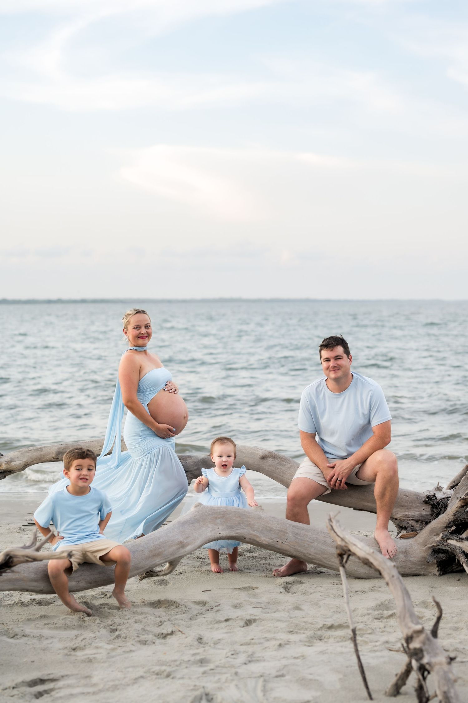 Pregnant family poses on driftwood at beach, with two young children, wearing matching blue outfits near ocean.