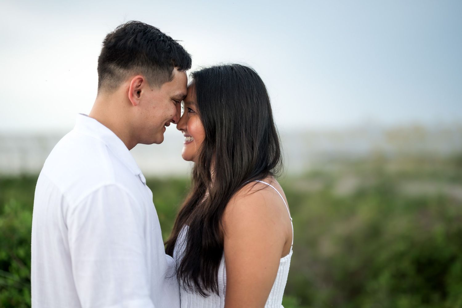 A romantic couple in white outfits touching foreheads and smiling outdoors near a beach.