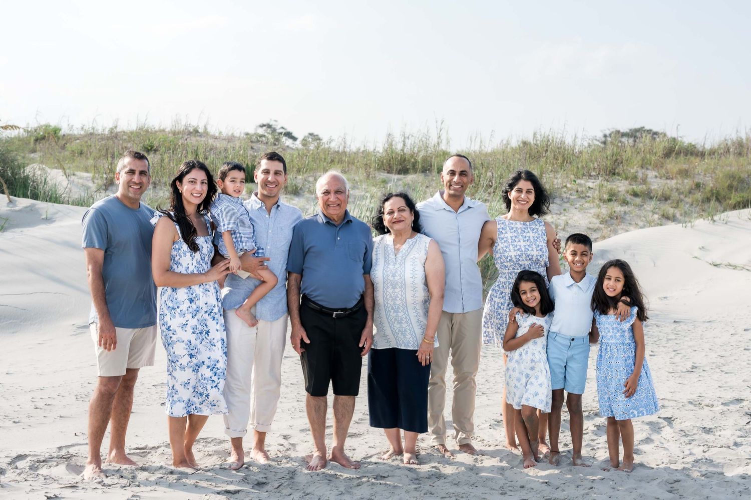 Multi-generational family posing together on a sandy beach, smiling in coordinated blue and white outfits.