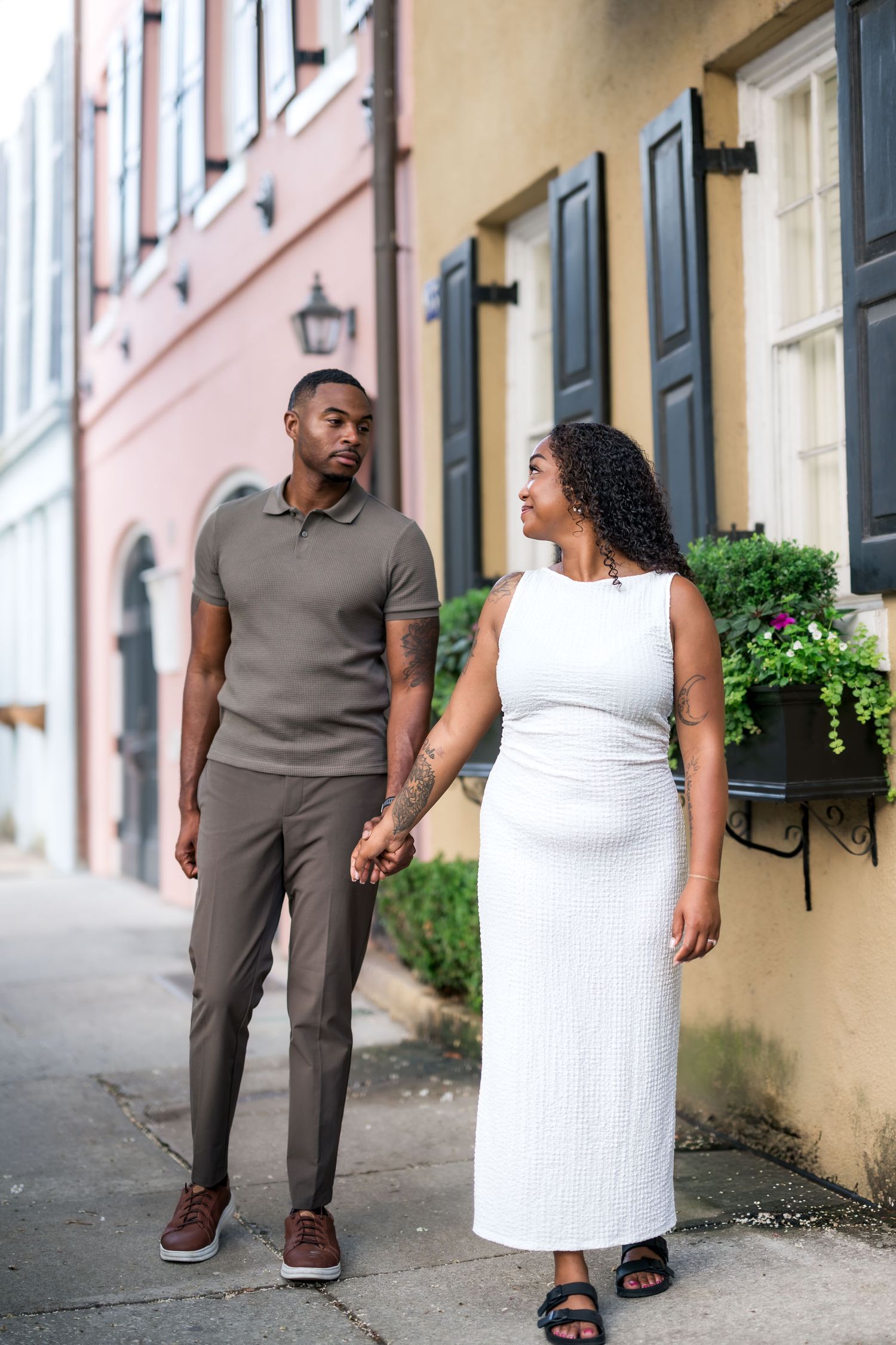 A couple holds hands while walking down a charming urban alley, woman in white dress, man in olive polo and brown trousers.