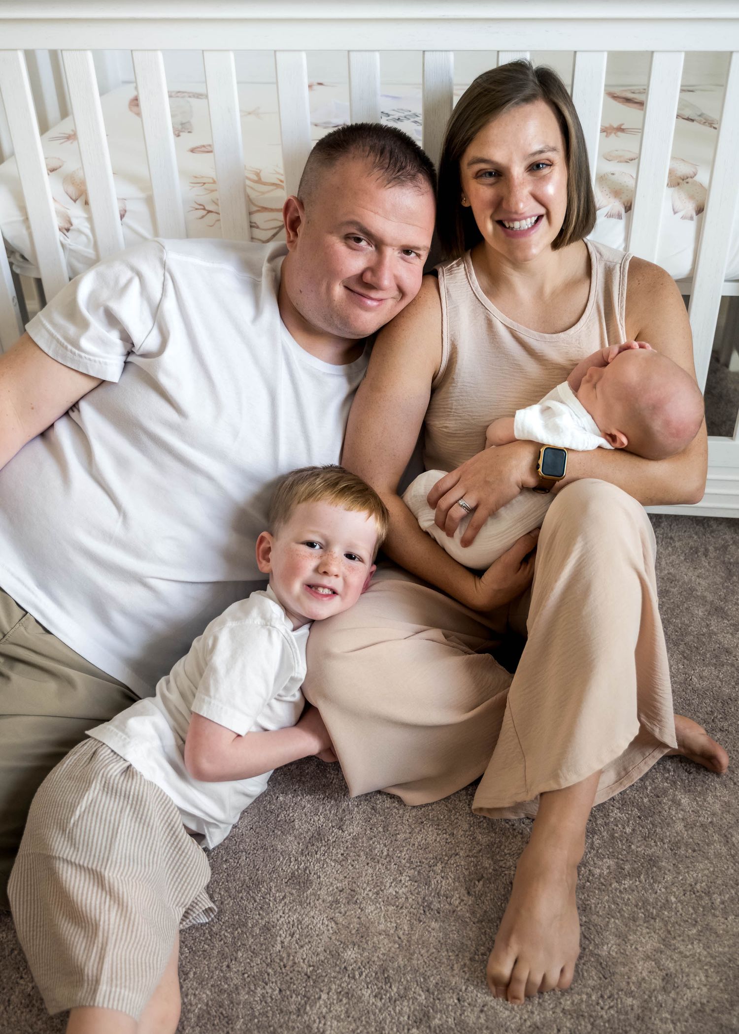 Happy family of four sitting by white crib, parents holding newborn with toddler smiling.