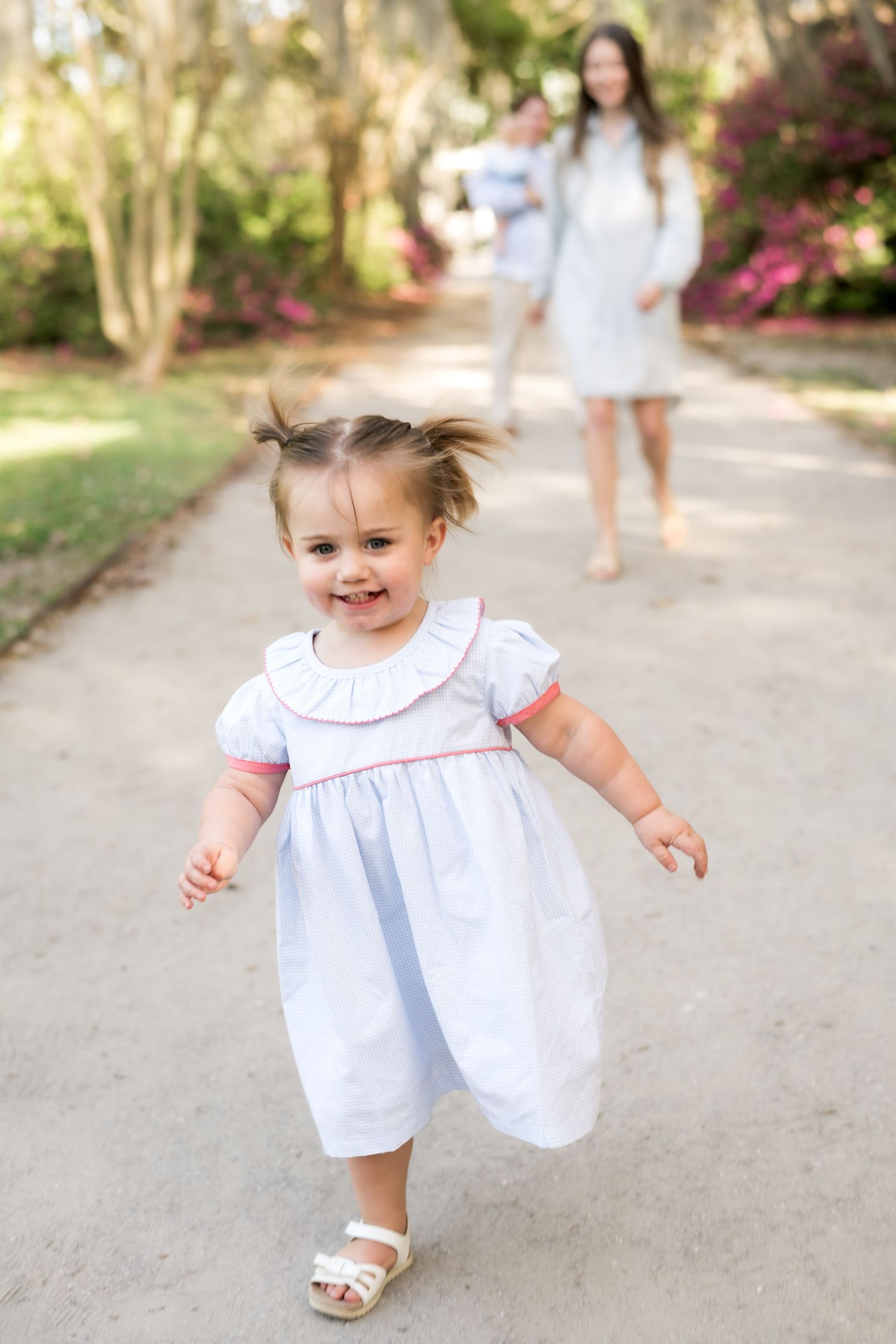 Toddler girl in white dress with pink trim runs on garden path while mother holds baby in background.