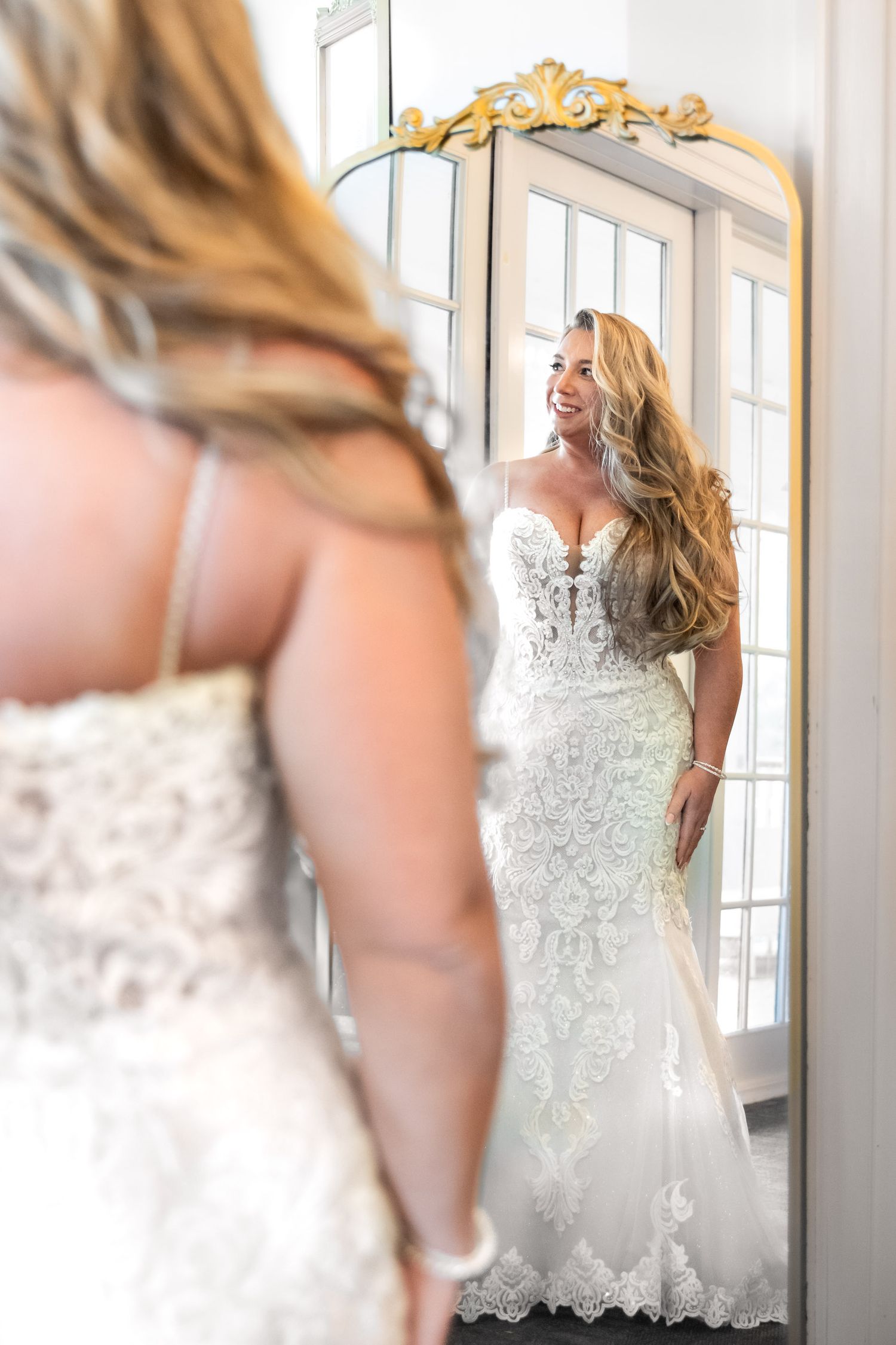 Dunes West Golf and River Club. Smiling bride in intricate lace mermaid wedding gown admiring her reflection in an ornate gold mirror.