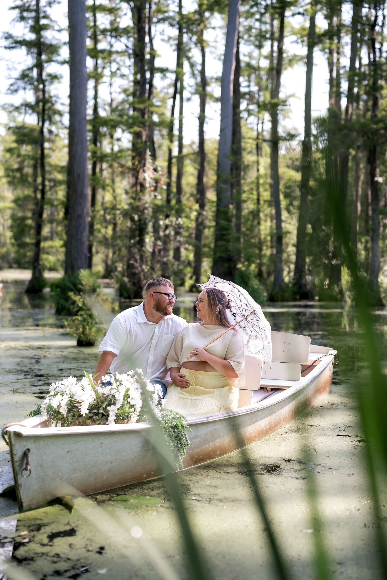 Couple in a white rowboat surrounded by cypress trees in a serene swamp, woman holding a parasol with flowers.