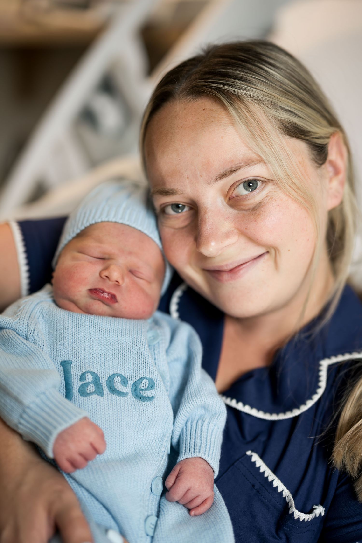 A smiling woman in blue scrubs holds a sleeping newborn dressed in a blue knit outfit embroidered with the name Lace.