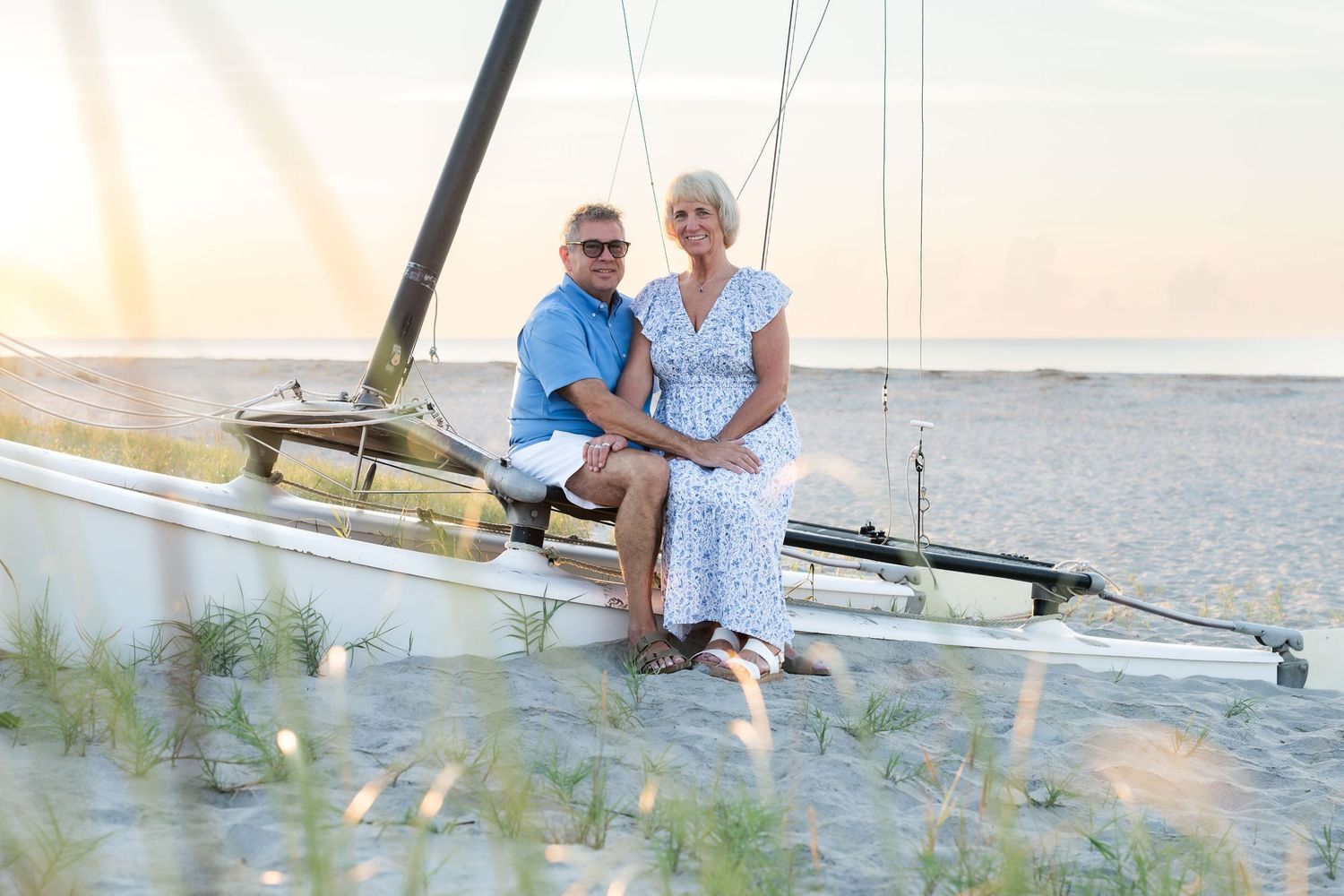 Couple sitting on a sailboat on a sandy beach at sunset, smiling together in casual summer attire.