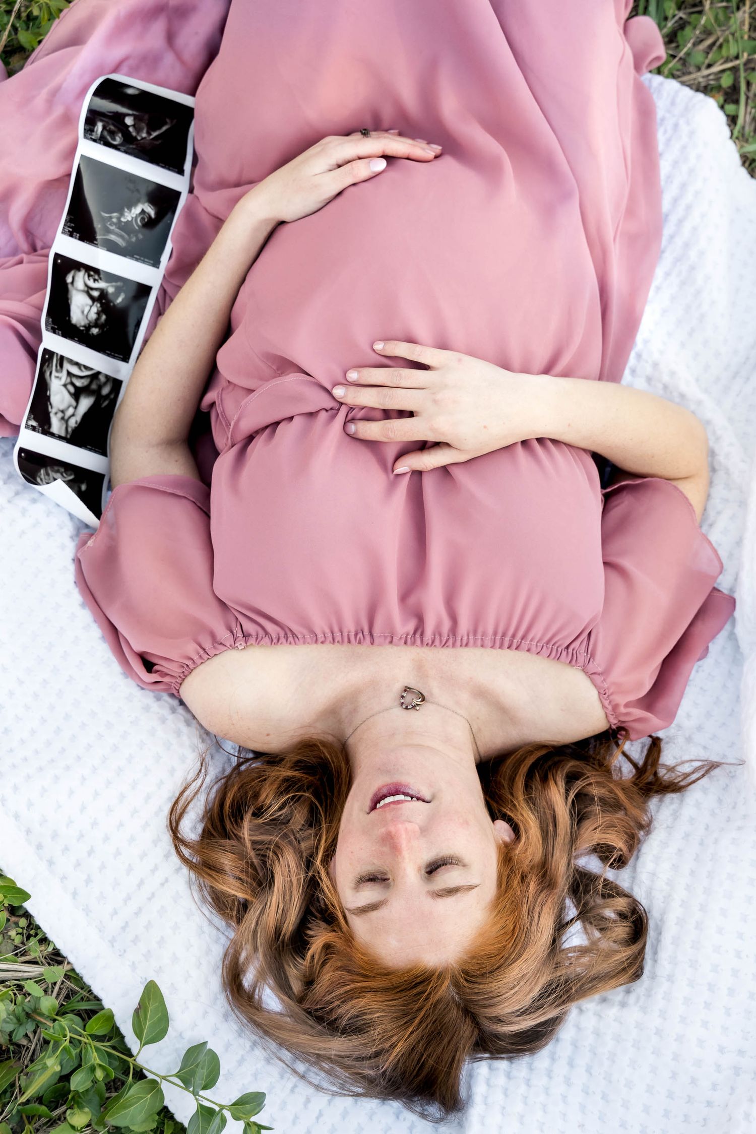 Moncks Corner.Pregnant woman in pink dress lies on white blanket outdoors holding baby bump with ultrasound photos beside her.