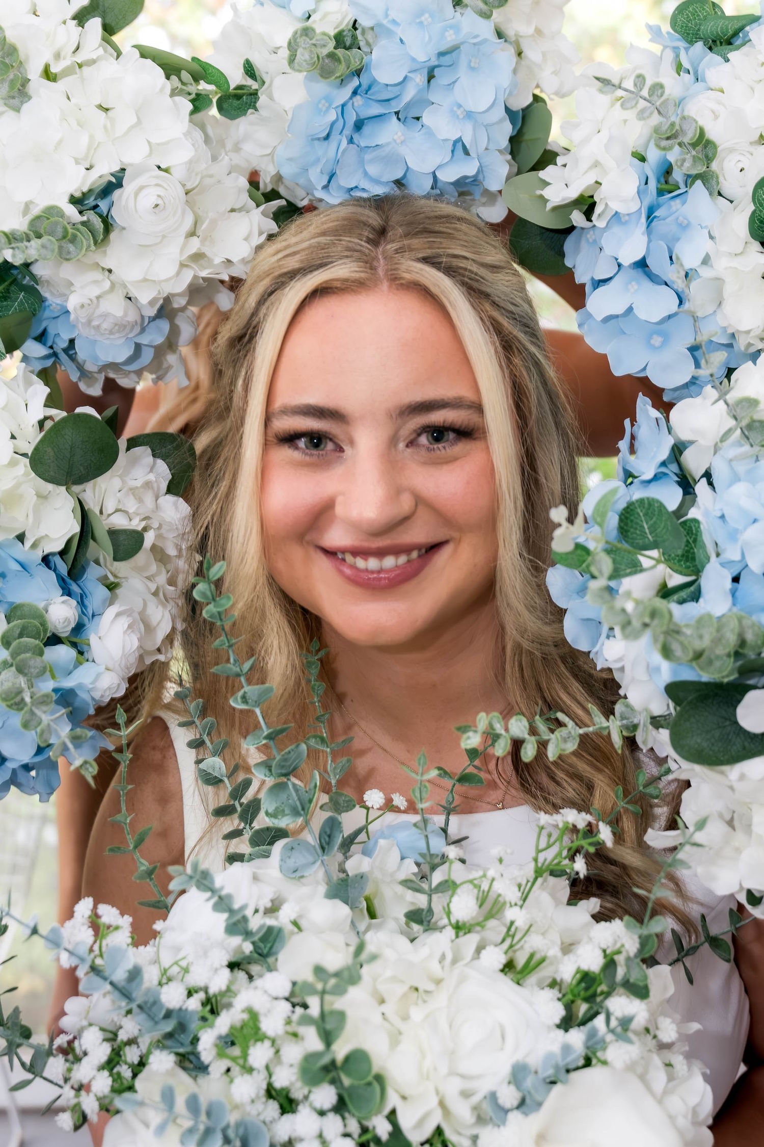 Smiling blonde bride framed by blue hydrangeas, white roses, and eucalyptus wedding arch flowers.