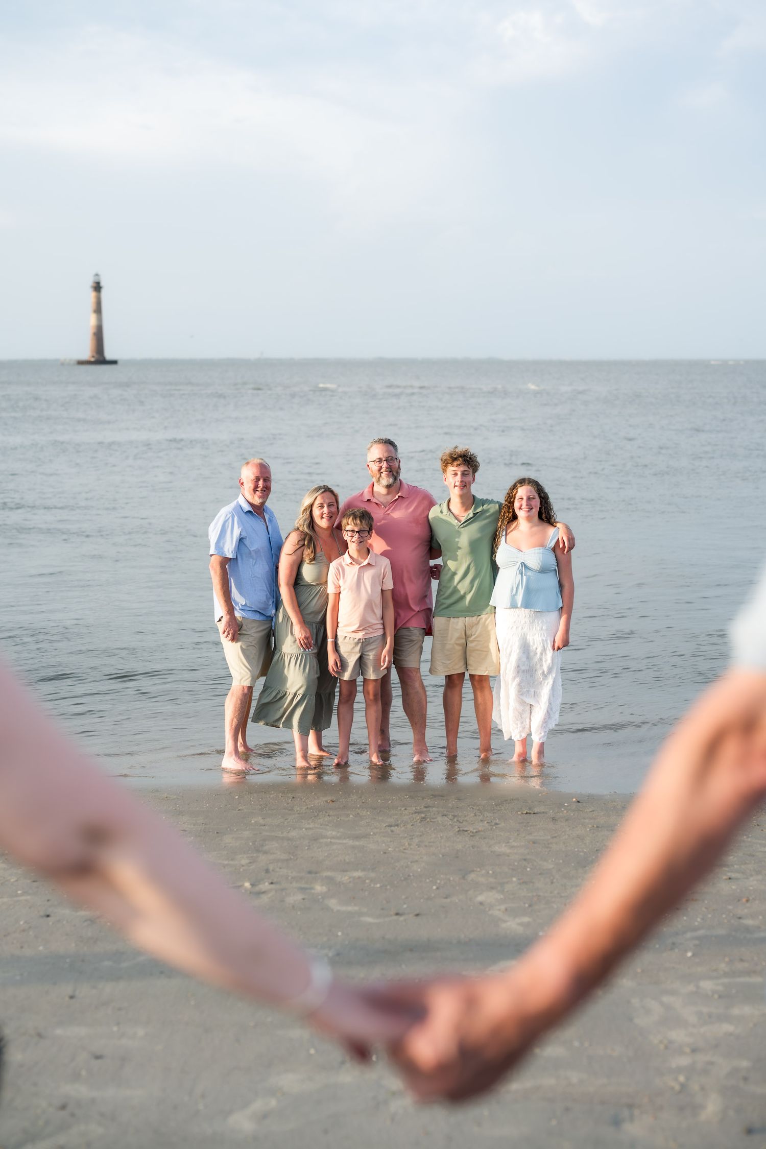 Multi-generational family smiling together on a beach with a lighthouse in the background on a sunny day.