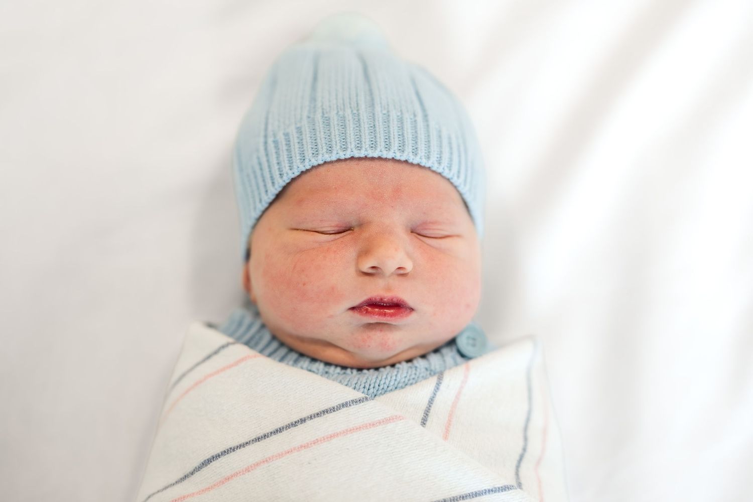 Peaceful newborn baby sleeping swaddled in white blanket wearing a soft blue knit hat.