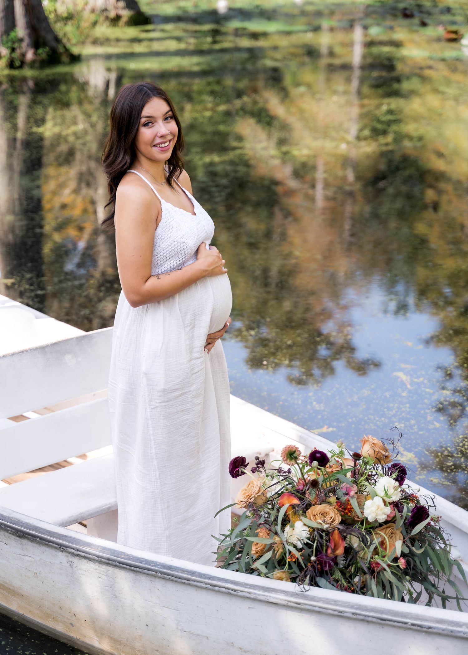 Glowing pregnant woman in white dress standing in rowboat with lush floral bouquet.