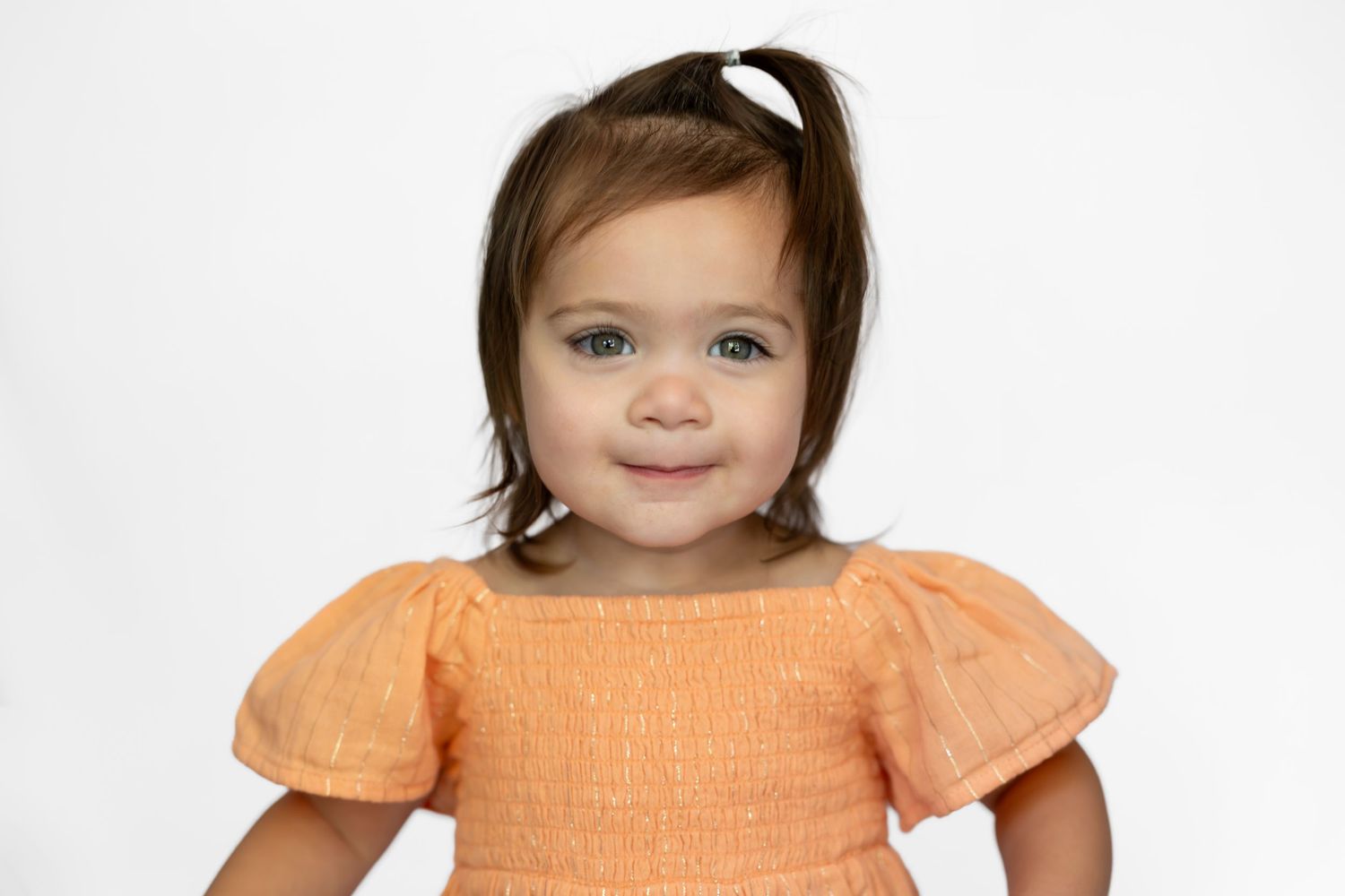 Smiling toddler girl with dark hair in orange dress posing for portrait photo on white background.