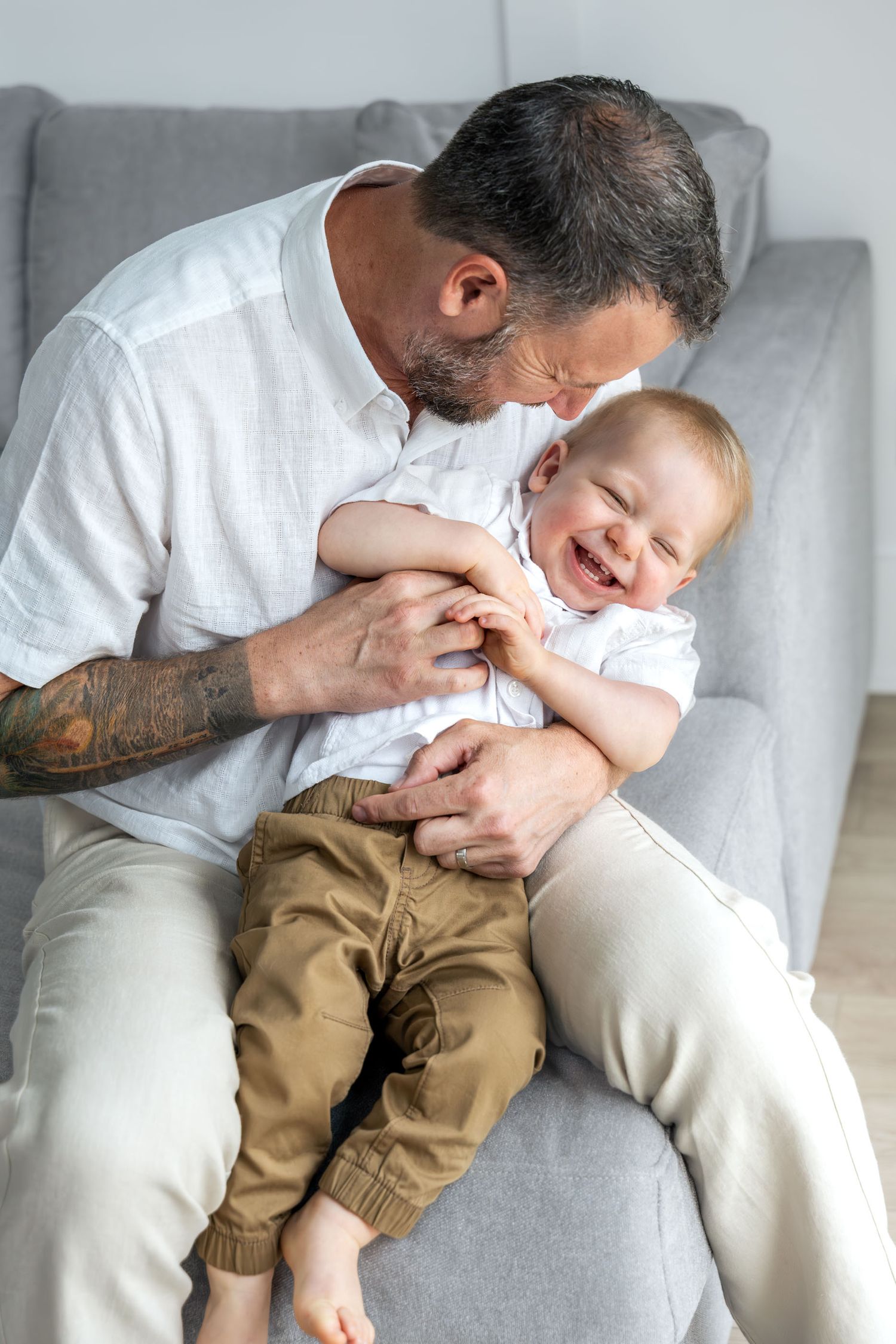 Tattooed father sits in gray chair laughing with his giggling baby boy in brown pants.