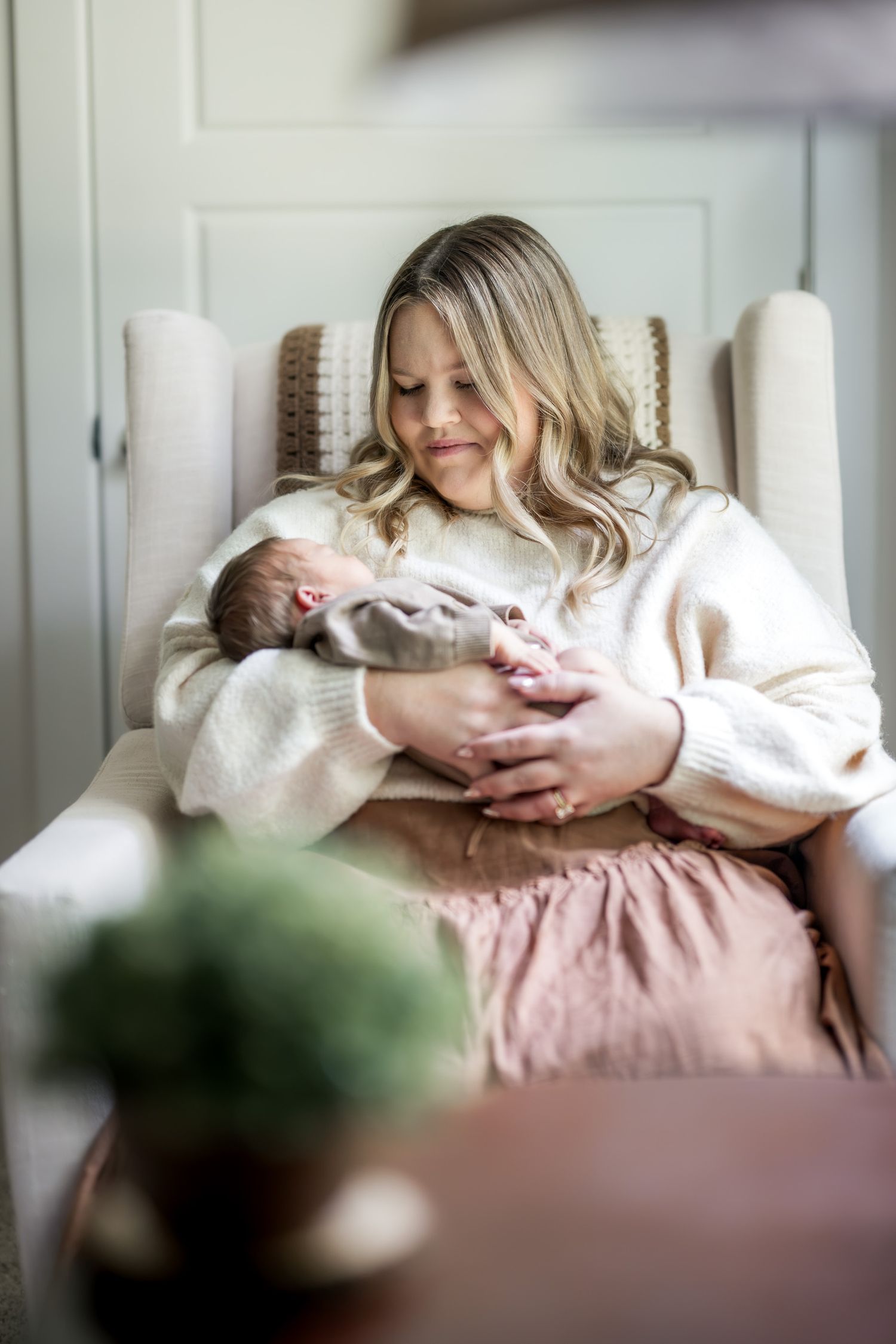 New mother gazes lovingly at sleeping newborn while seated in a nursery chair.