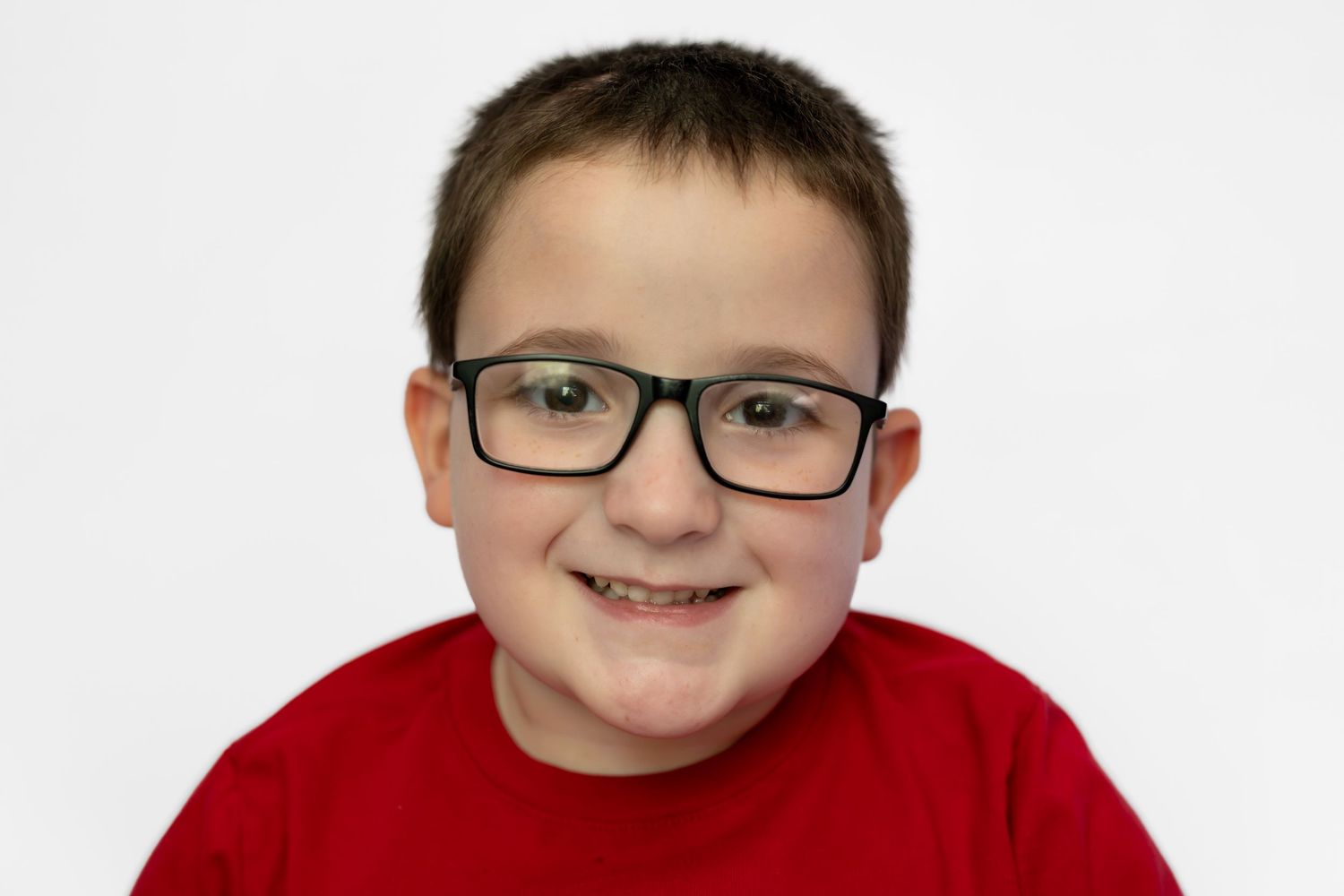 Smiling young boy with black glasses wearing a red t-shirt against a white background.