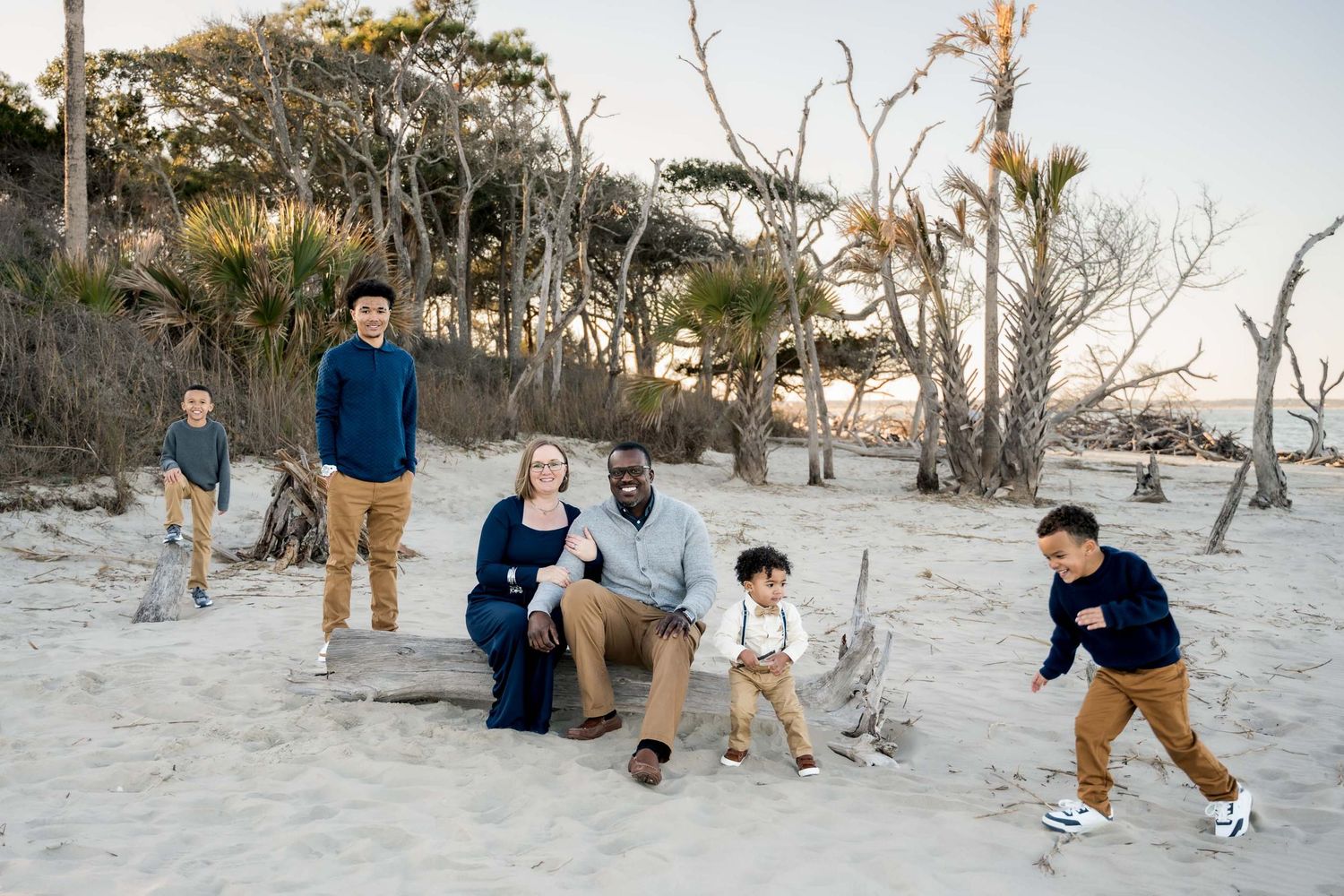 Family of six relaxes on driftwood at a scenic beach with bare trees and golden sunset light.