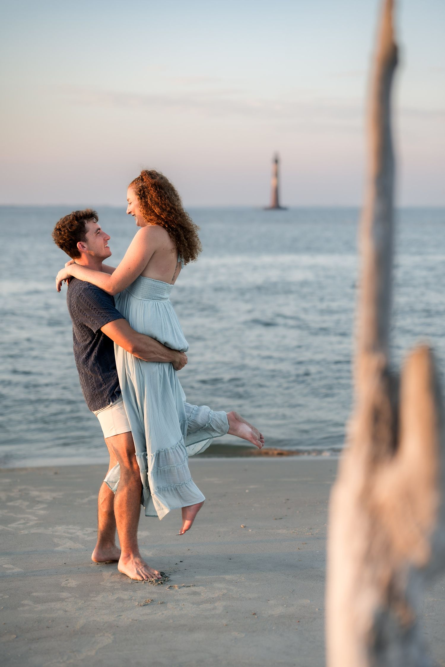 Couple embracing on beach at sunset with lighthouse in background, man lifting woman in blue dress.