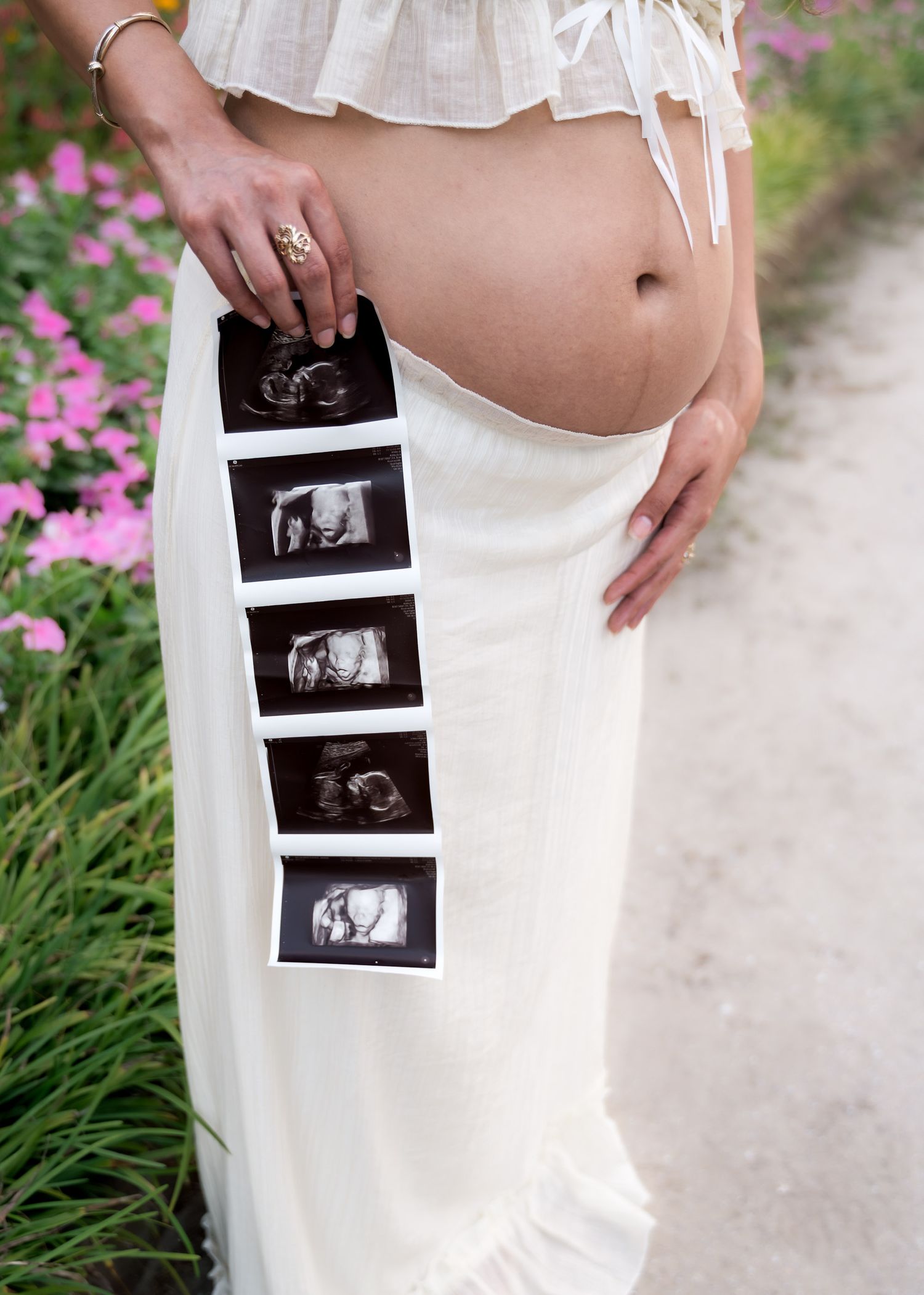 Pregnant woman in white skirt holding ultrasound photos beside her baby bump, surrounded by pink flowers.