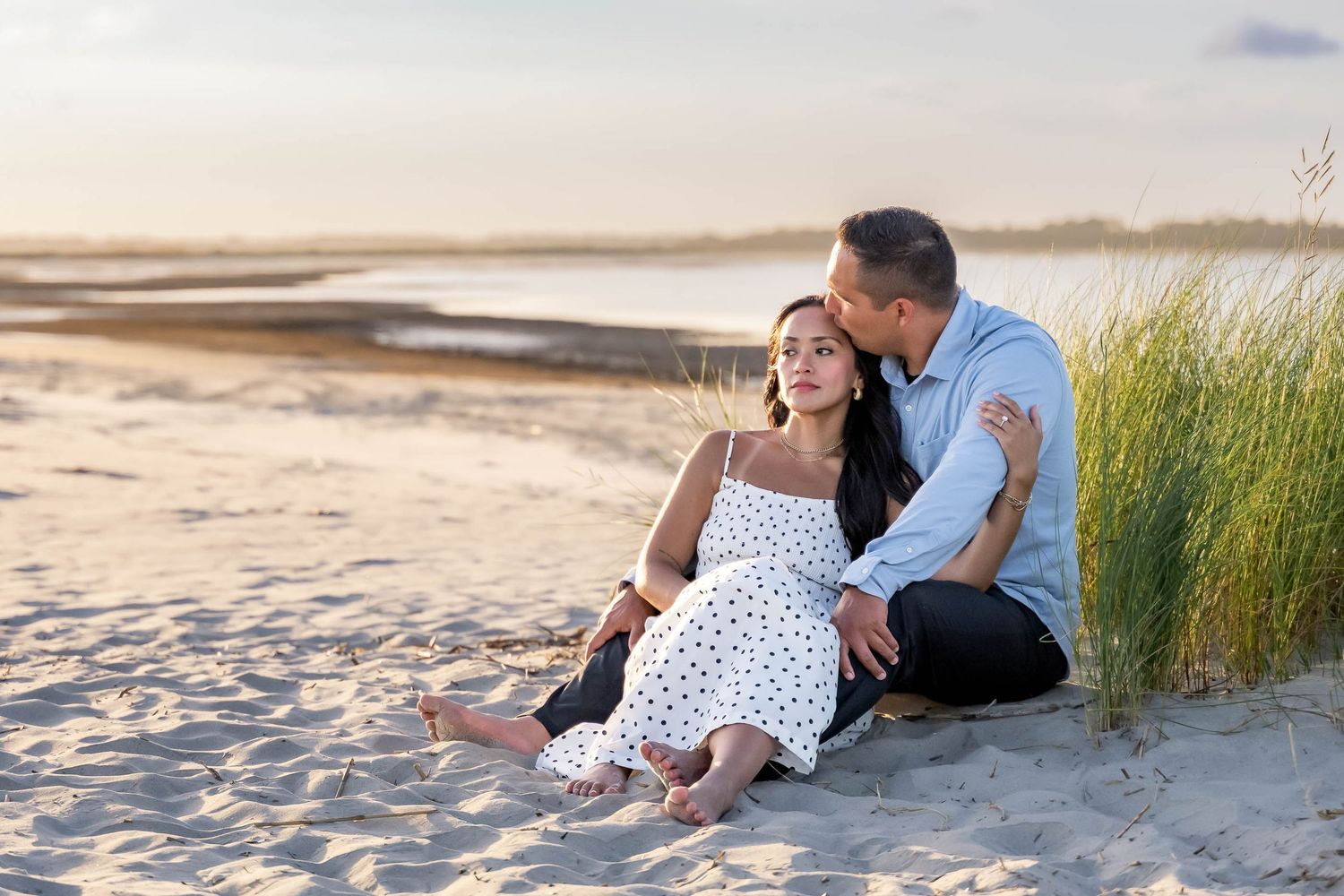 Folly Beach. Couple sitting on beach sand at sunset, man kissing woman's head, she wears white polka dot dress.