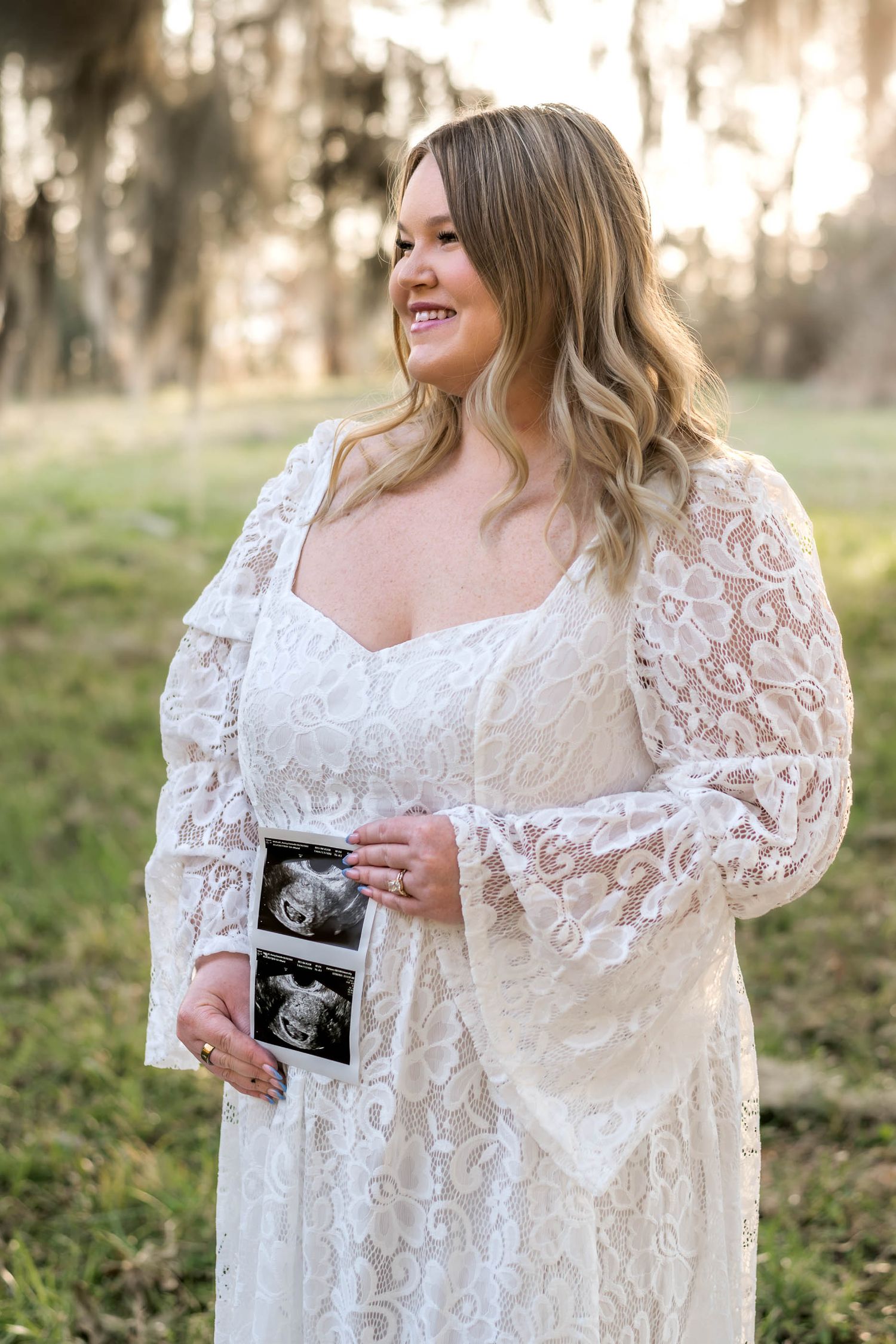 Moncks Corner.Smiling pregnant woman in white lace dress holding ultrasound photos outdoors in soft light.