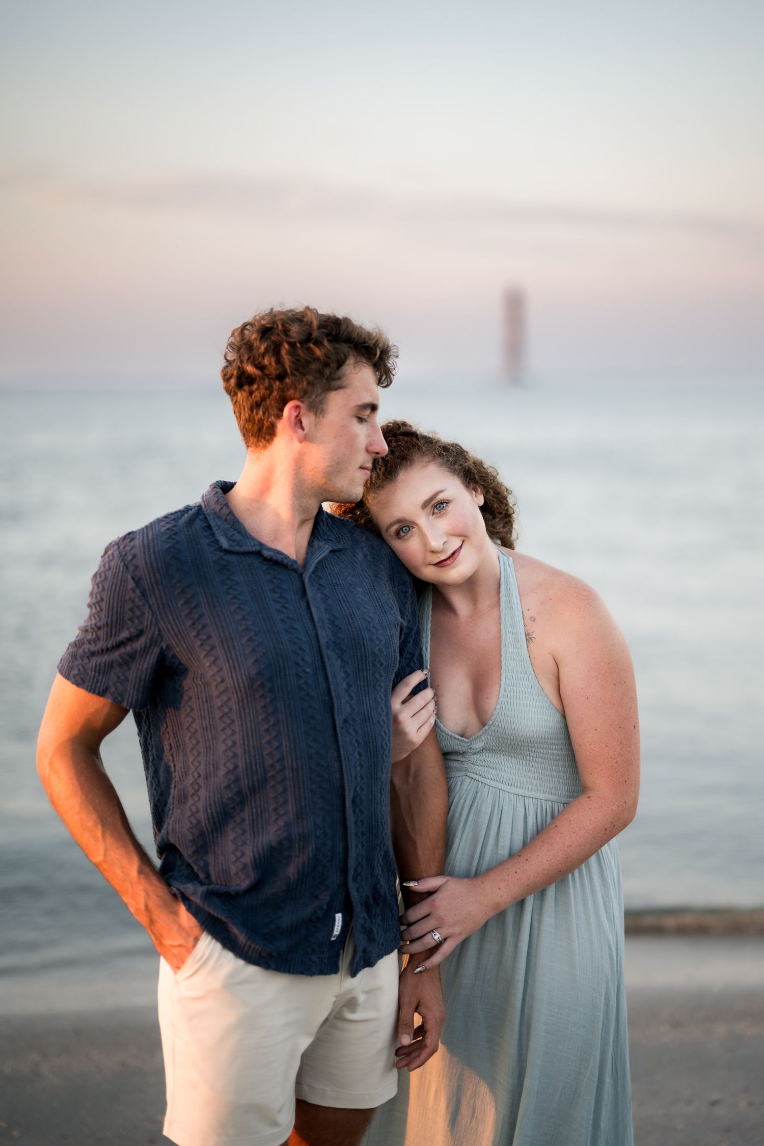 Couple embracing at sunset on a beach, woman in sage halter dress leaning on mans shoulder, lighthouse in background.
