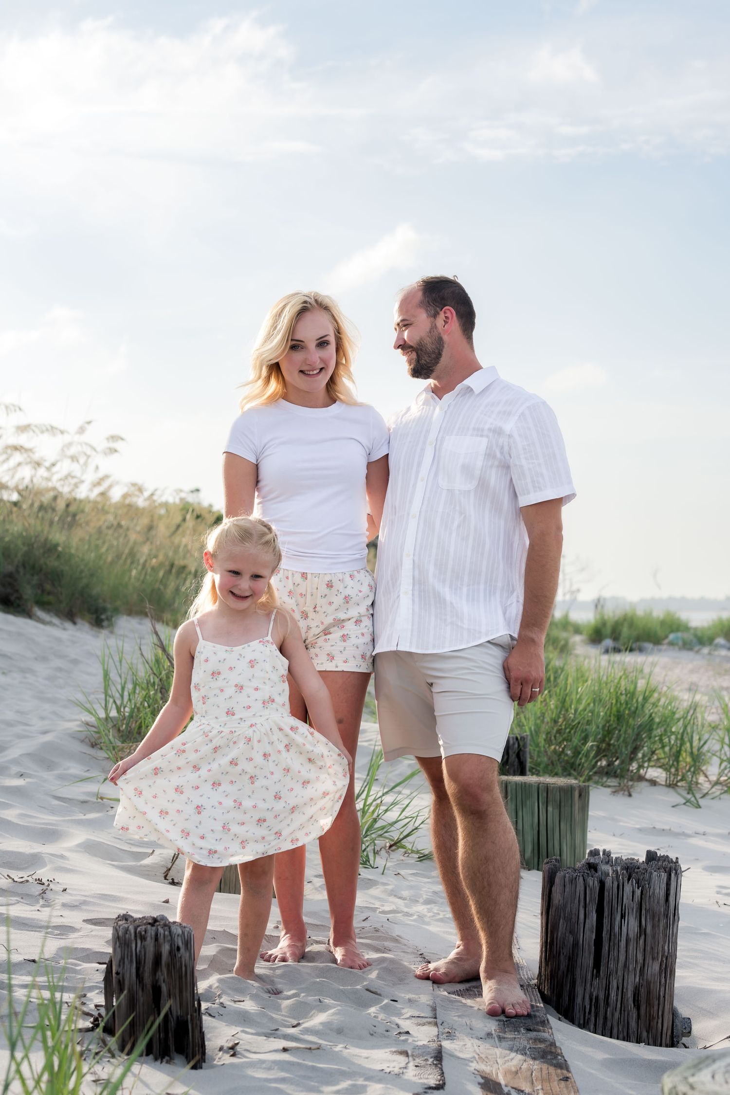 Family of three enjoying a sunny beach day, with a little girl twirling in a floral dress on sandy dunes.
