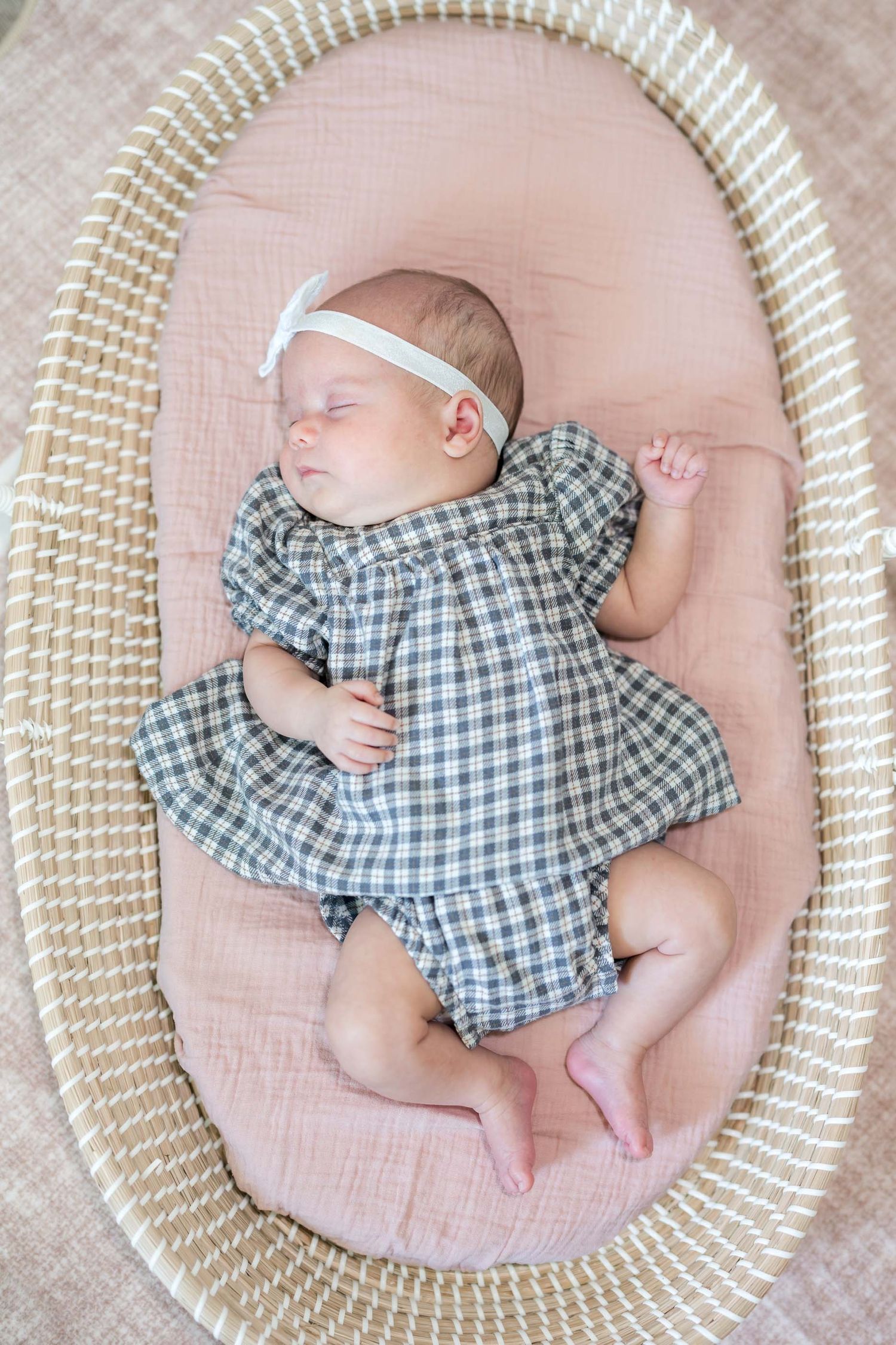 Sleeping newborn baby girl in plaid dress and white bow headband lying in a wicker Moses basket with pink liner.