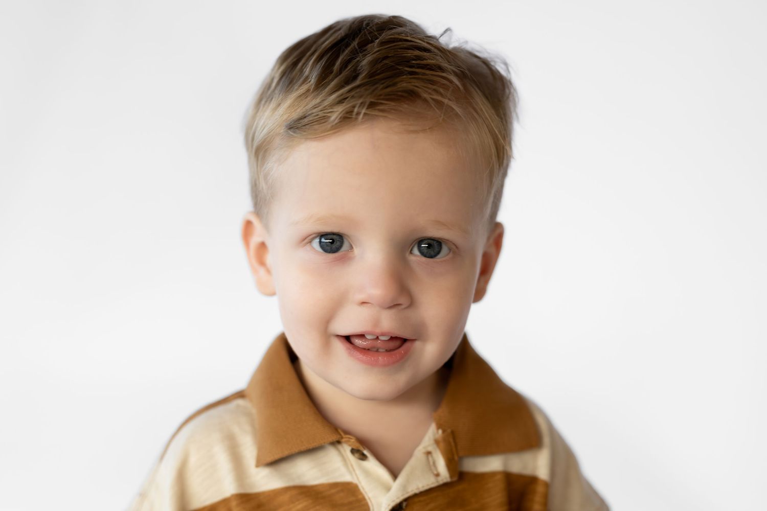 Smiling toddler boy with blonde hair and blue eyes wearing a brown striped polo shirt against white background.