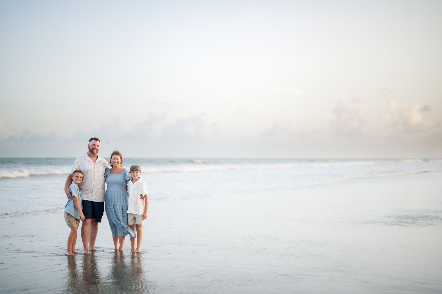 Family of four smiling together on a sandy beach at sunset with gentle ocean waves in the background.