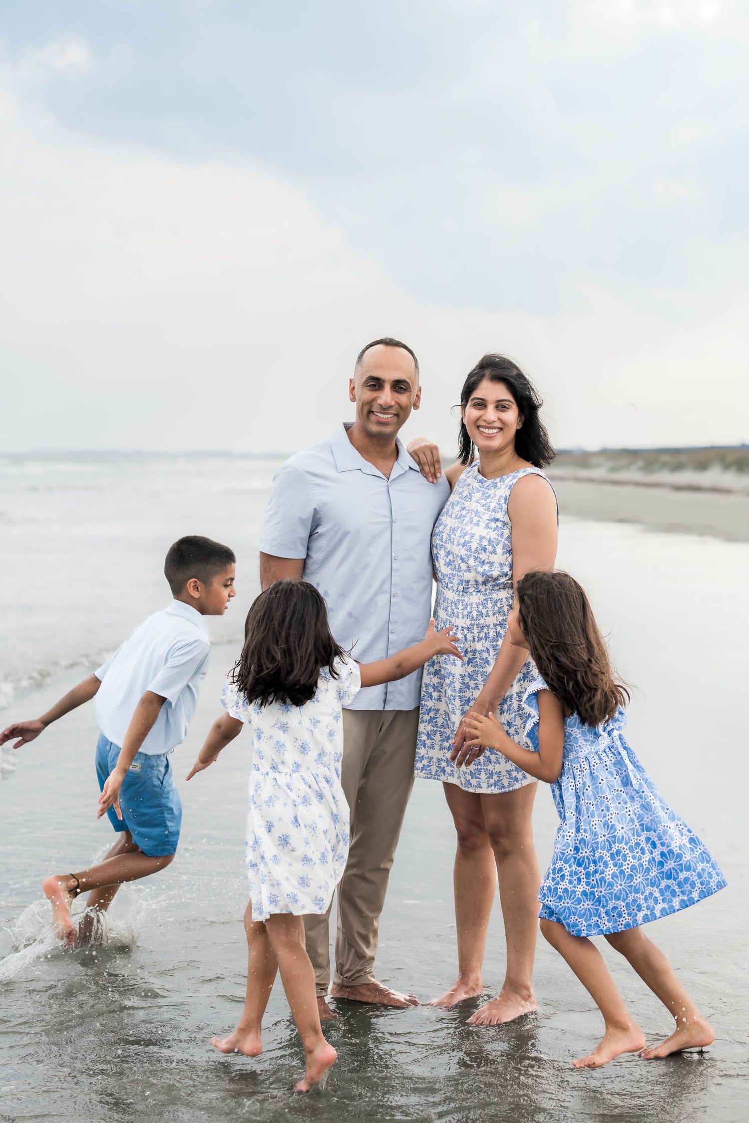Family of five smiling together on a sandy beach, parents embracing while three children play in the ocean waves.