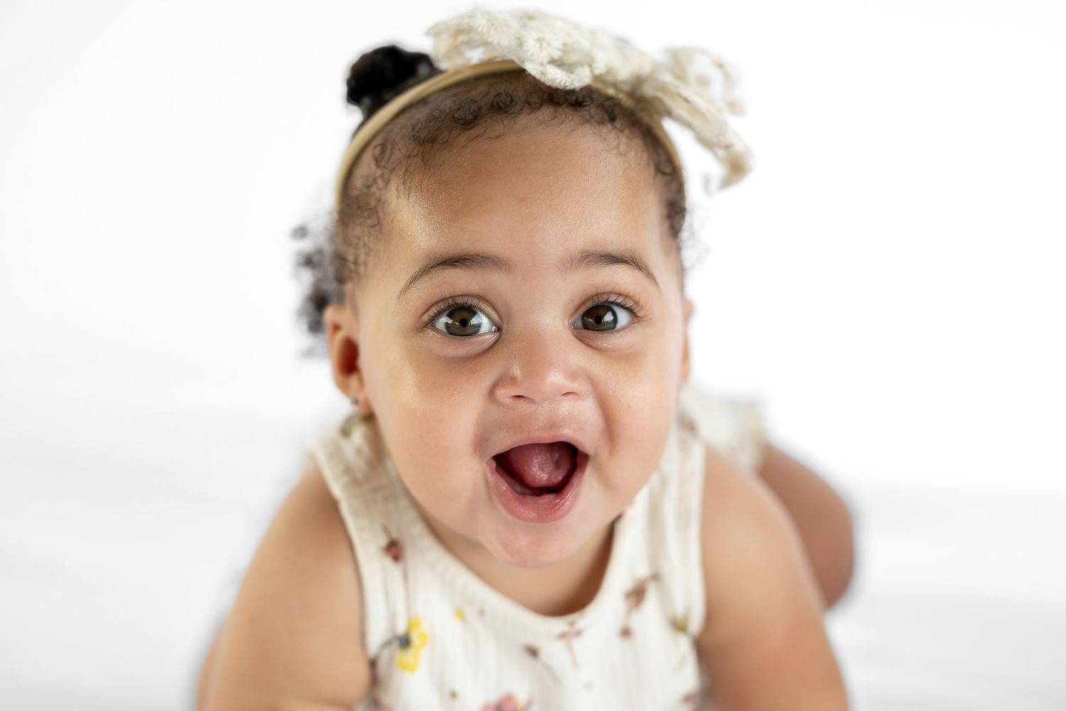 Adorable baby girl with a bow headband smiling with mouth open, wearing a white floral outfit.
